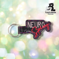 Front view of a black glitter vinyl keychain die-cut to the design outline, with "NEURO" embroidered in pale pink block lettering above "Spicy" in red-coral script, finished with pink and purple double-stitched edge detailing and a black plastic snap closure with silver split ring.