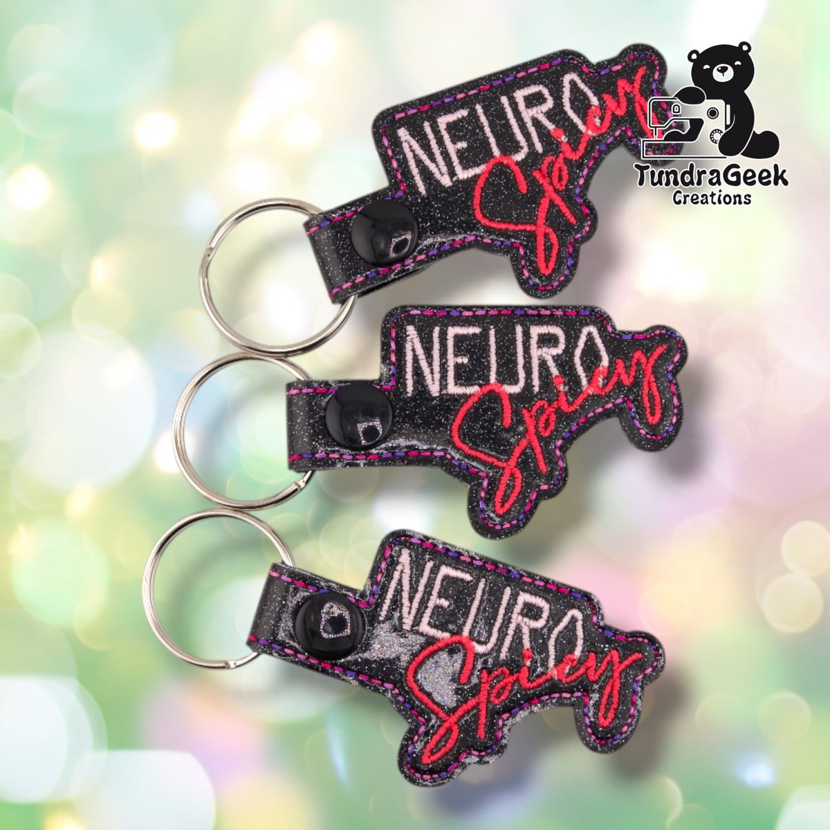 Three of the same keychain shown together at slightly different angles, each in black glitter vinyl with "NEURO" in pale pink block letters and "Spicy" in red-coral script, matching pink and purple stitched edges and black plastic snap closures.