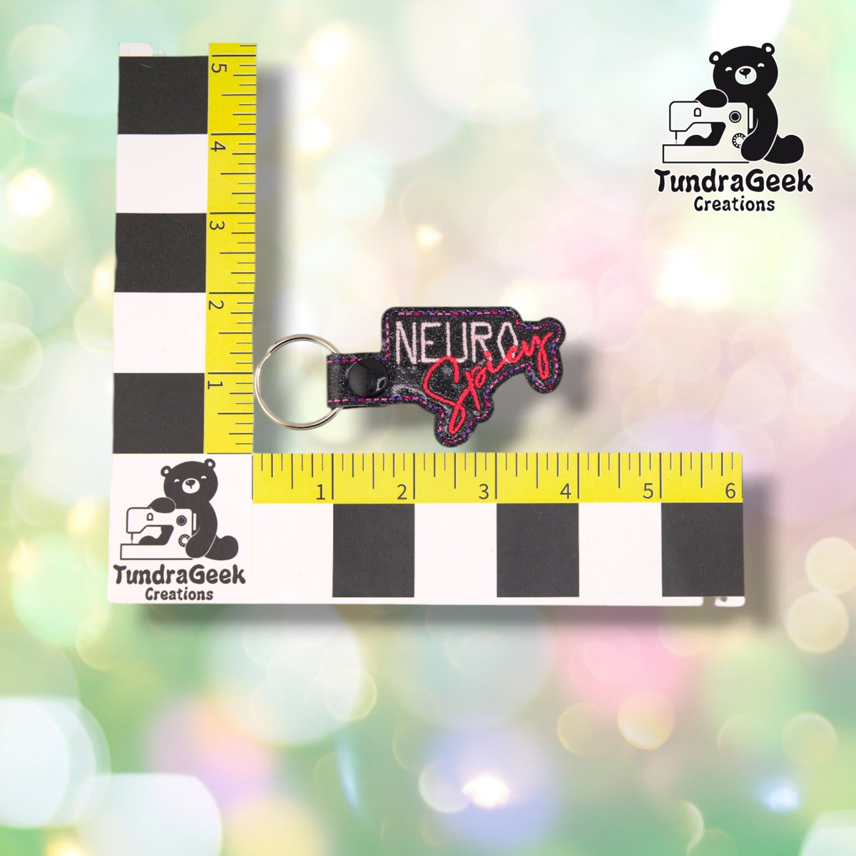 Keychain shown against a black-and-white checkered background with a yellow ruler marked in inches, measuring approximately 4 inches wide by 2 inches tall including hardware.  ---