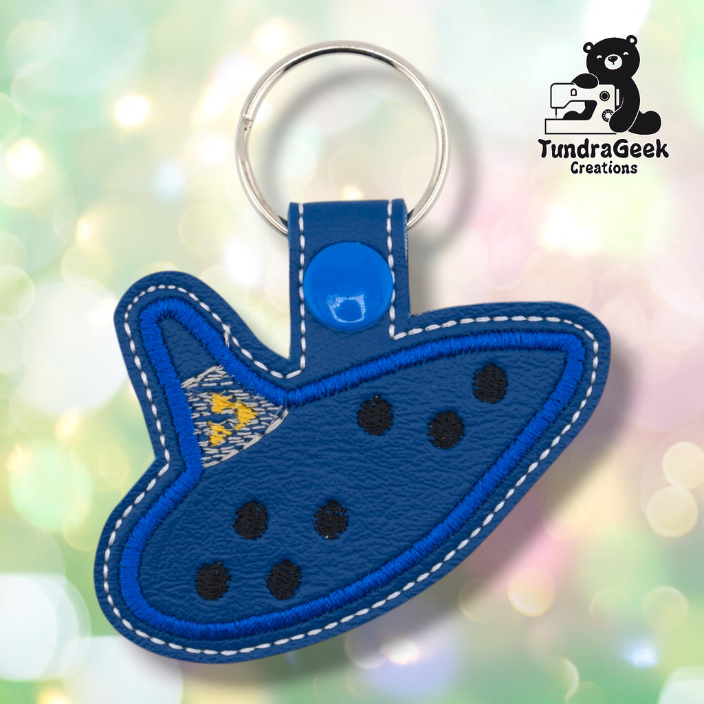Blue faux leather keychain shaped like an ocarina, styled after the iconic instrument from a classic fantasy video game. Bright blue embroidered border and eight finger holes are visible on the dark navy body; a small cluster of yellow stars is embroidered near the mouthpiece. Blue snap closure and silver key ring at top. TundraGeek Creations logo in upper right corner.