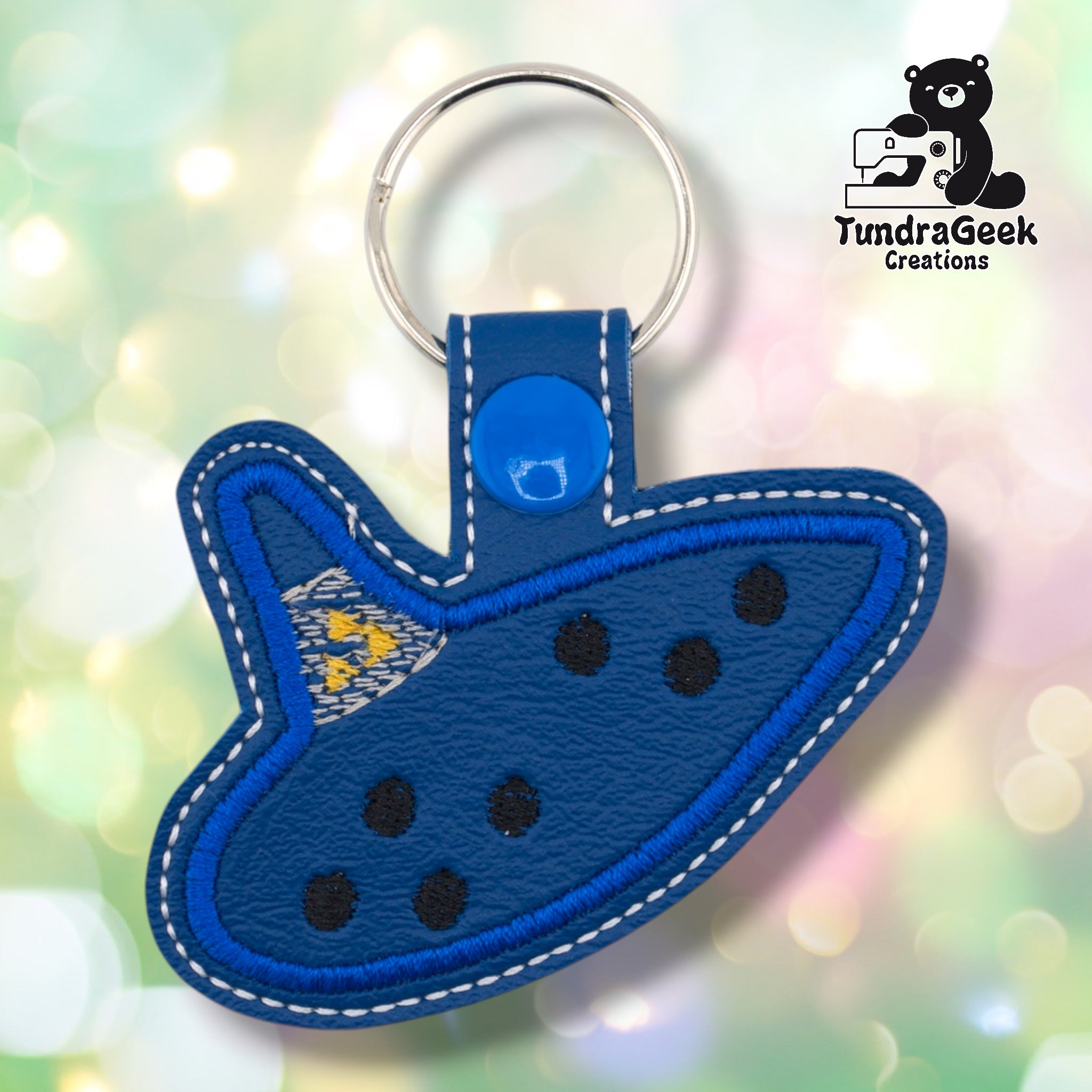 Blue faux leather keychain shaped like an ocarina, styled after the iconic instrument from a classic fantasy video game. Bright blue embroidered border and eight finger holes are visible on the dark navy body; a small cluster of yellow stars is embroidered near the mouthpiece. Blue snap closure and silver key ring at top. TundraGeek Creations logo in upper right corner.