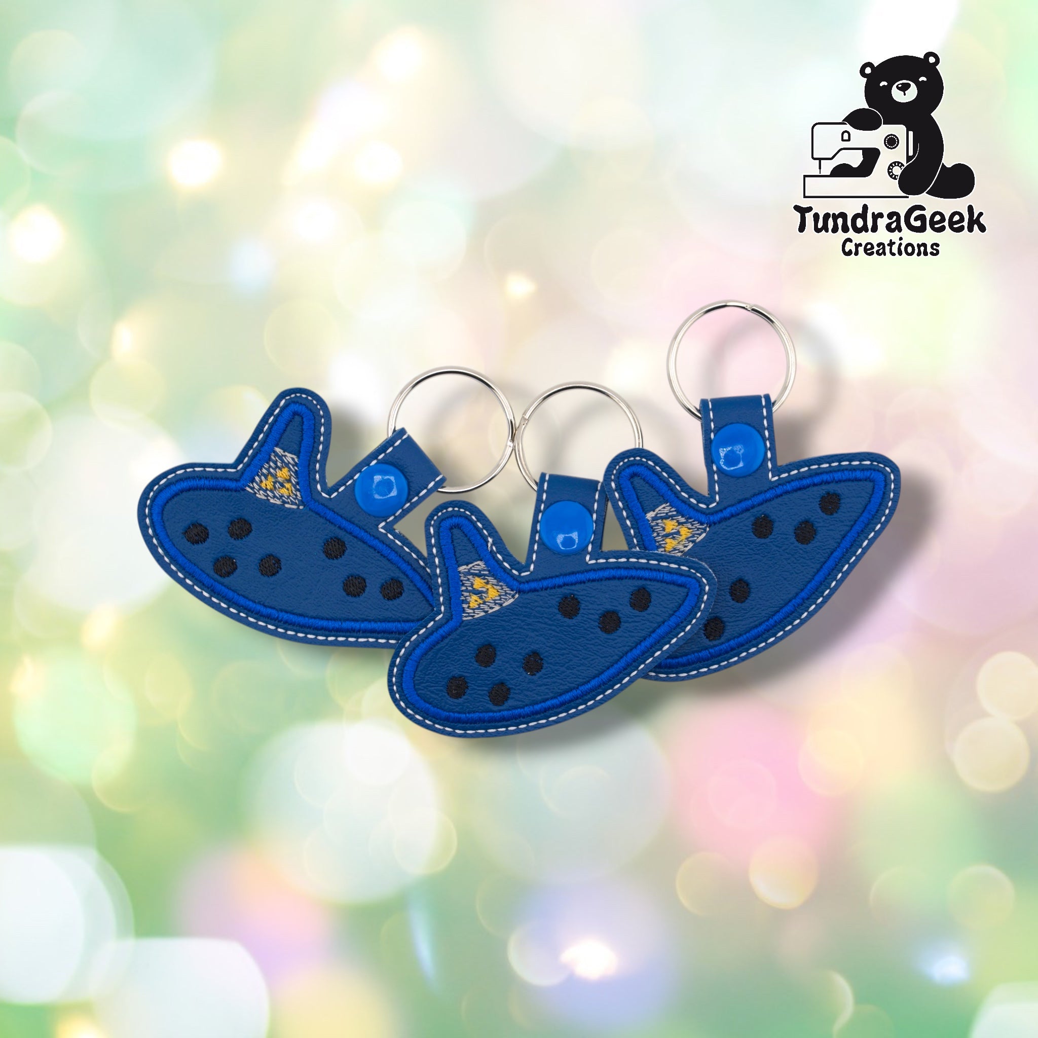 Three ocarina keychains arranged together on a pastel bokeh background, showing the design from slightly varied angles. All three feature the same navy and blue colorway with yellow star detail.