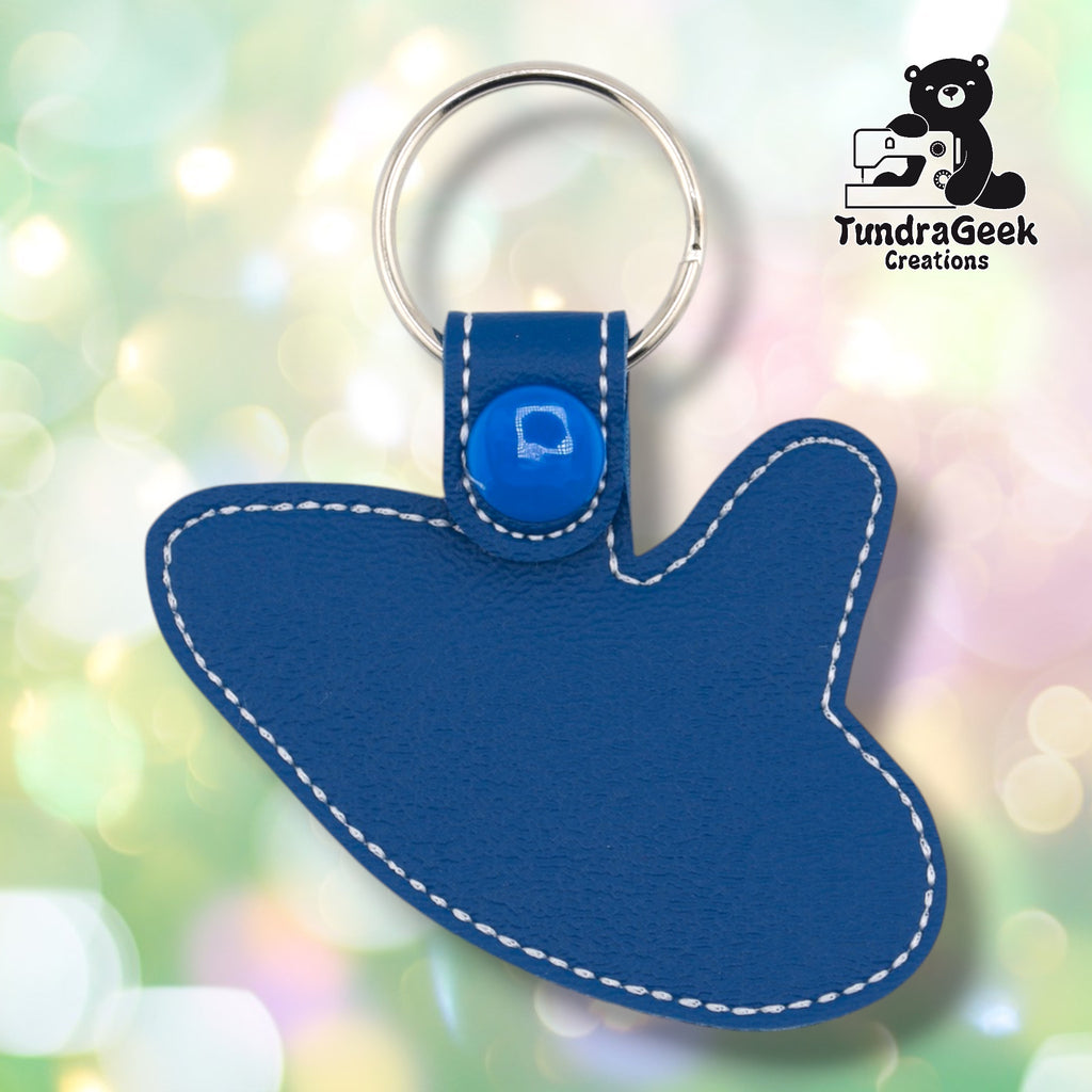 Plain back of the ocarina keychain showing blue faux leather, white outline stitching following the instrument shape, and blue snap closure. No embroidery on reverse.
