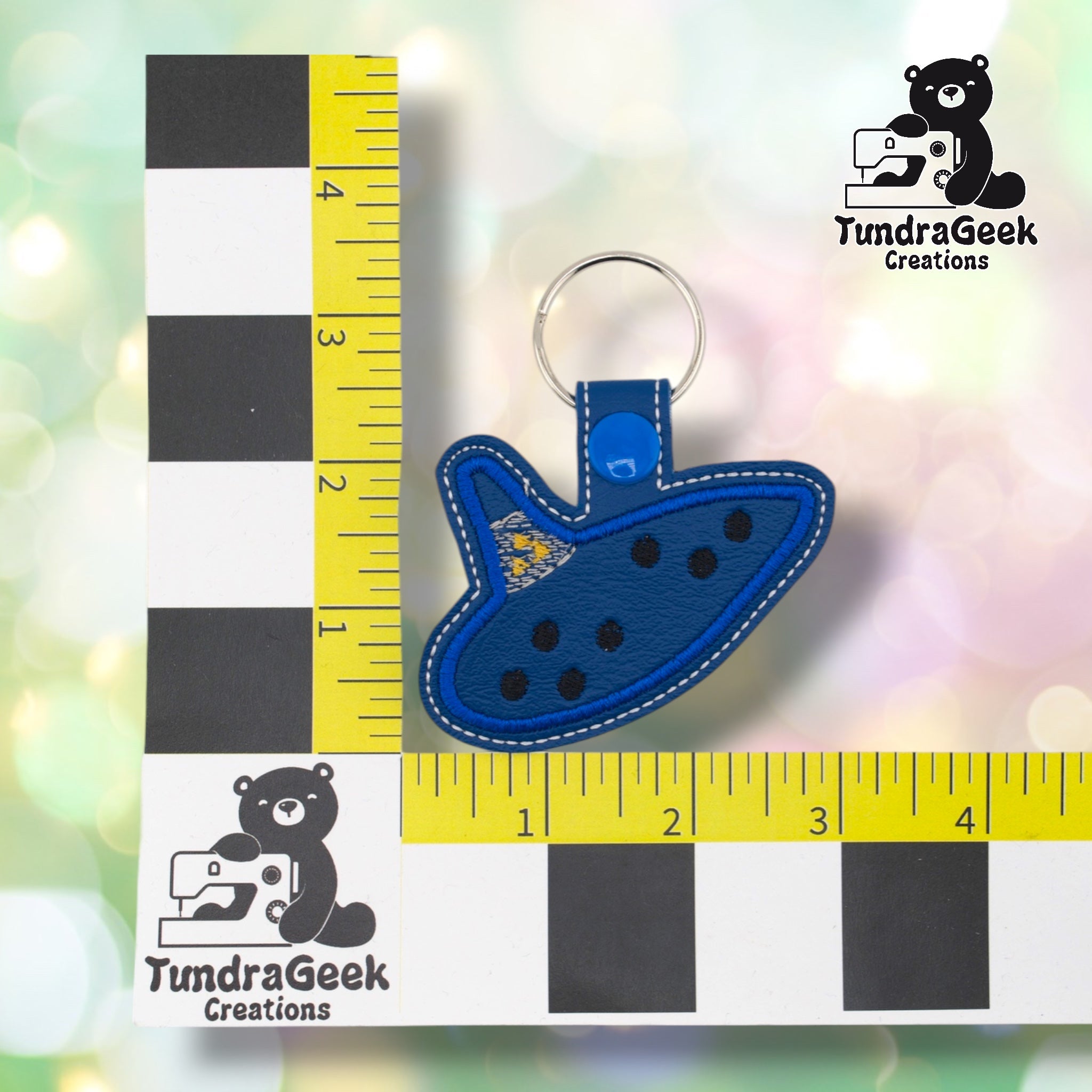 Ocarina keychain positioned against a TundraGeek Creations branded ruler card showing yellow tape measure. Keychain body measures approximately 3 inches wide by 2 inches tall, not including hardware.