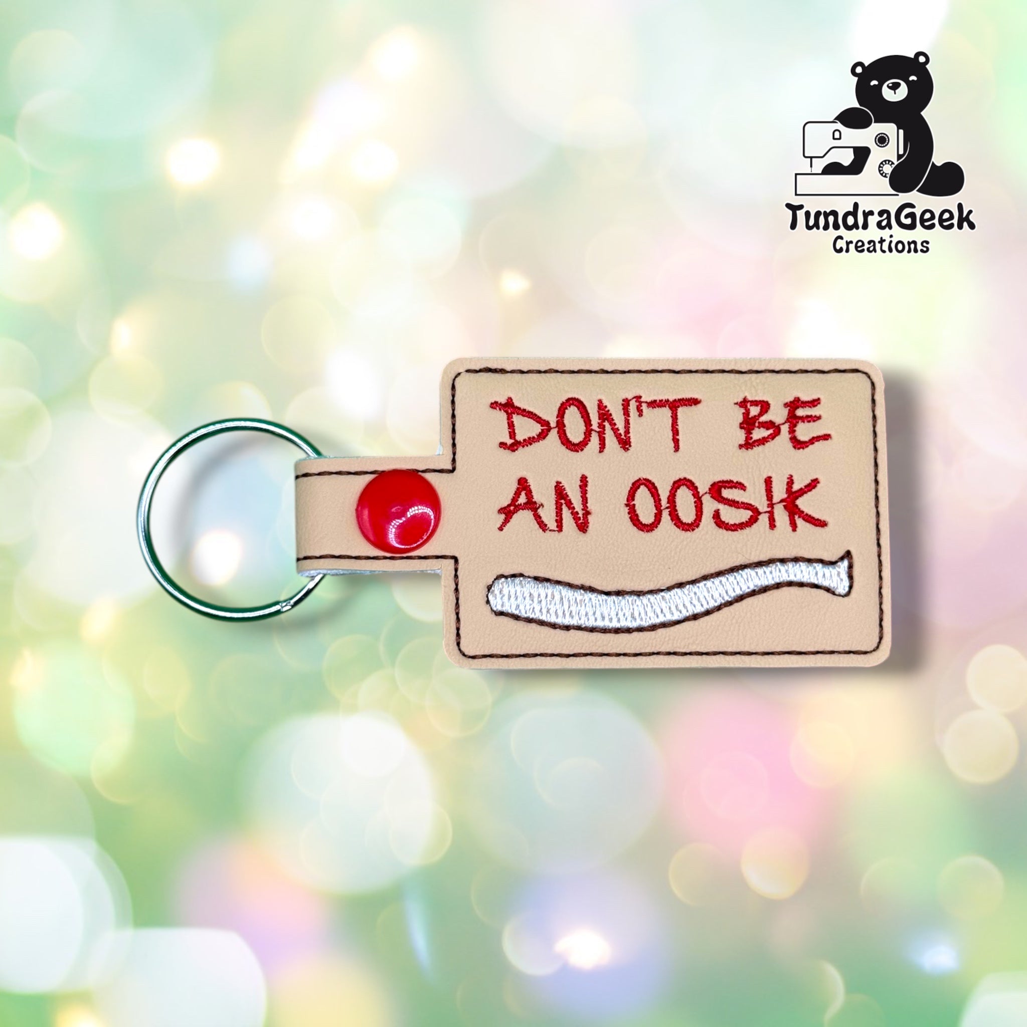 Front view of a rectangular tan vinyl keychain reading "DON'T BE AN OOSIK" in red embroidered script, with a white embroidered oosik bone illustration stitched below the text, finished with a red plastic snap closure.