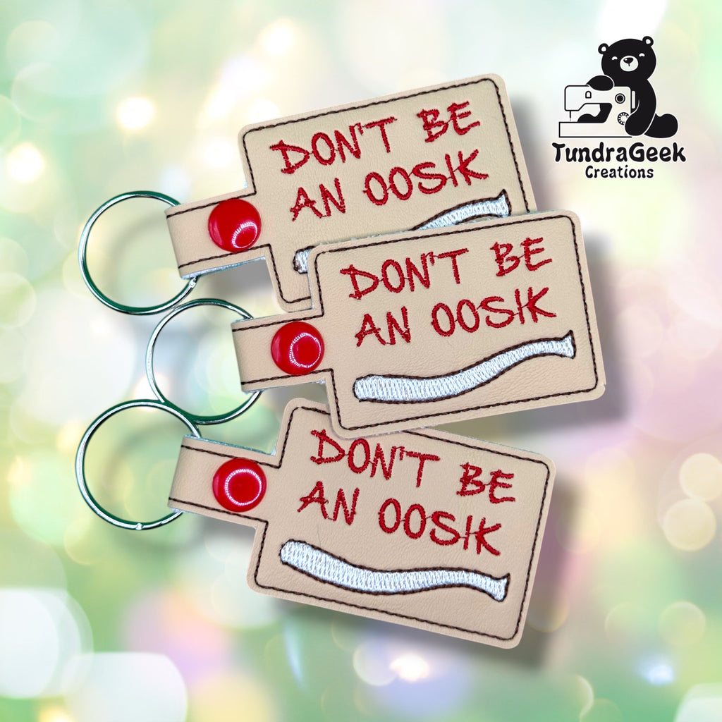 Three of the same keychain fanned out together, each tan vinyl with "DON'T BE AN OOSIK" embroidered in red above a white oosik bone illustration, red plastic snap closures, and split rings.