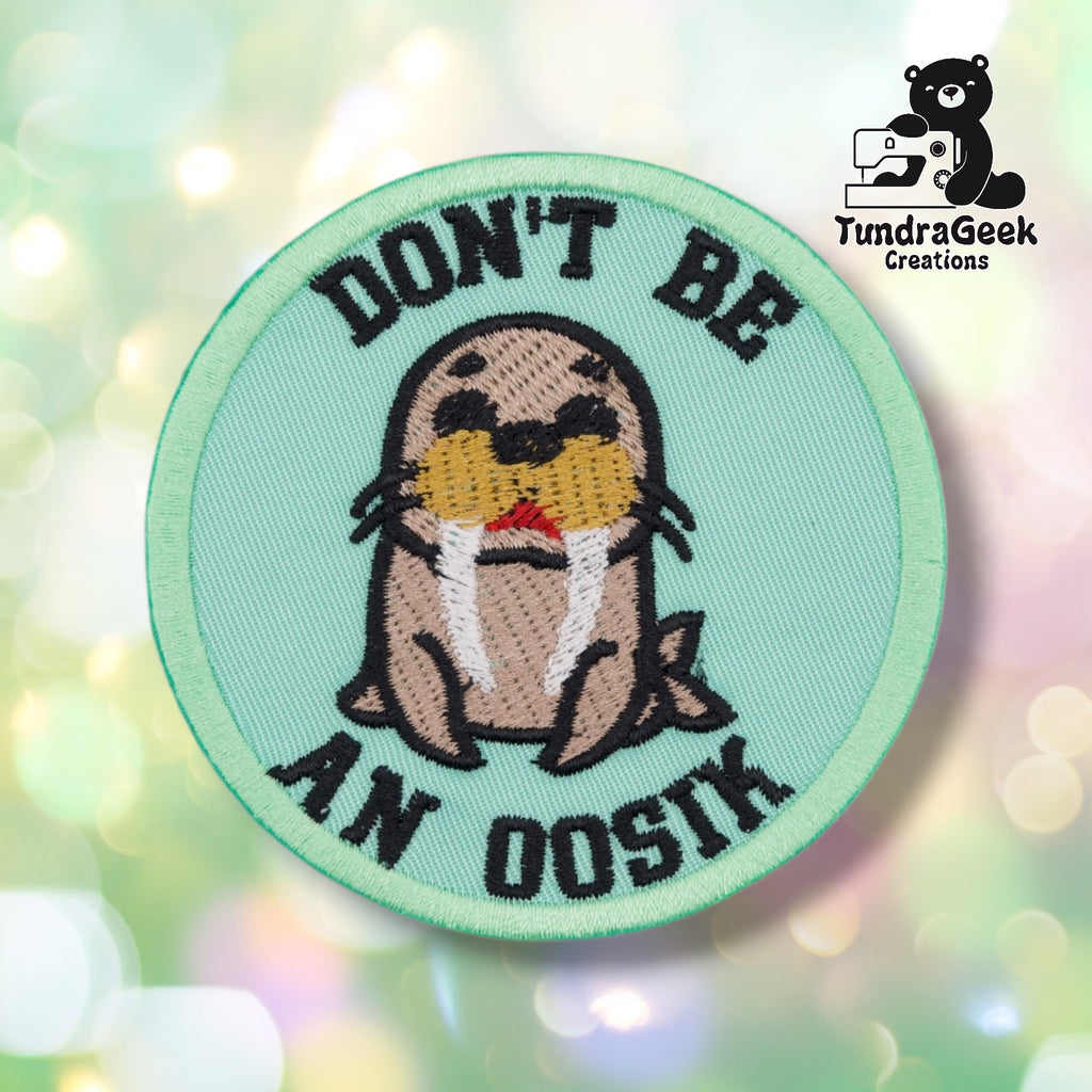 A single round patch centered on a soft pastel bokeh background, shown flat for a clear view of the design. A cartoon walrus with dark eyes, white tusks, and a red mouth sits centered on a mint green background, with "DON'T BE" arched across the top and "AN OOSIK" arched across the bottom in bold black lettering. Mint green border edging.