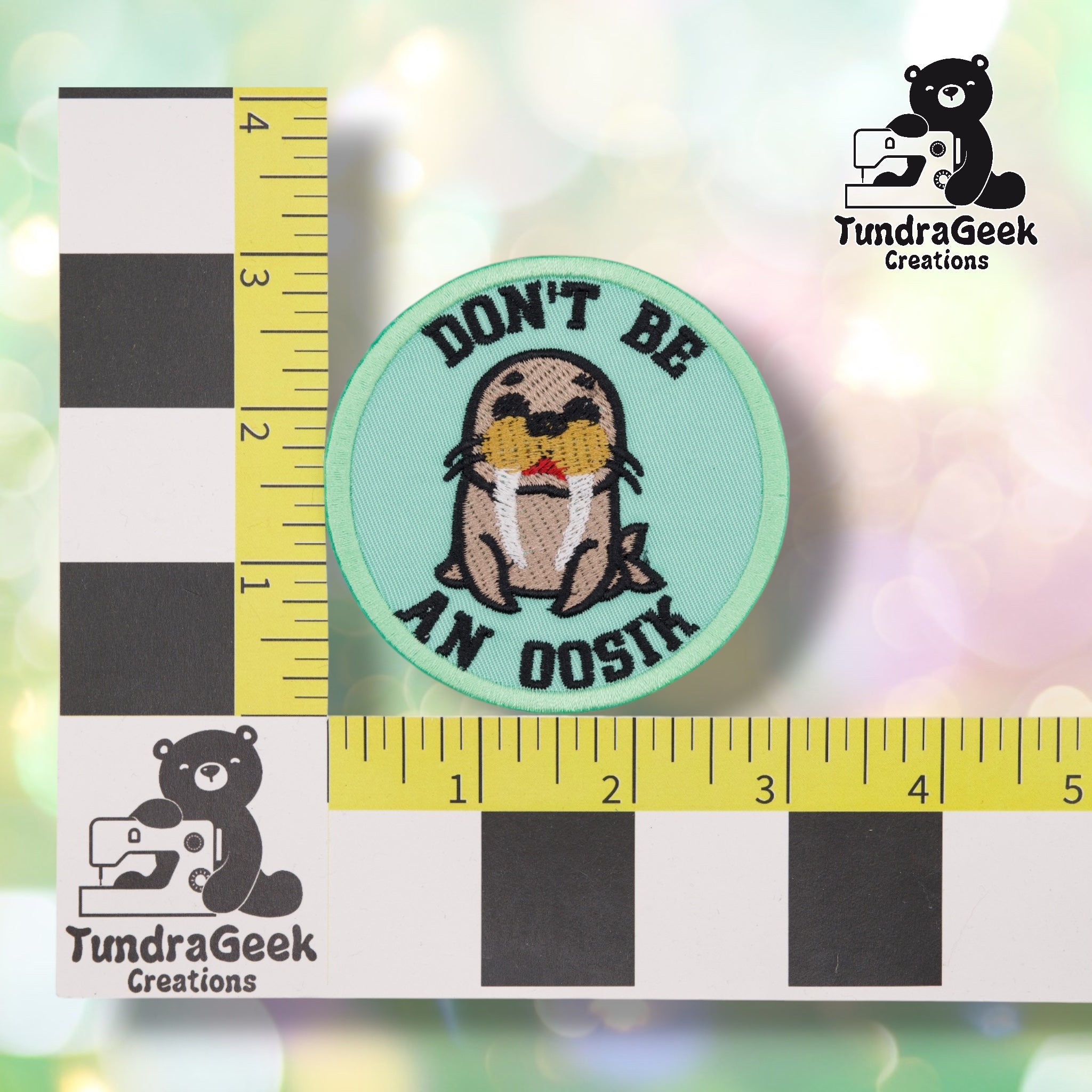 The patch shown against a black-and-white checkered background with yellow rulers for scale, measuring 3 inches in diameter. The design shows a cartoon walrus with dark eyes, white tusks, and a red mouth, mint green background and border, with "DON'T BE" and "AN OOSIK" arched in bold black lettering above and below.