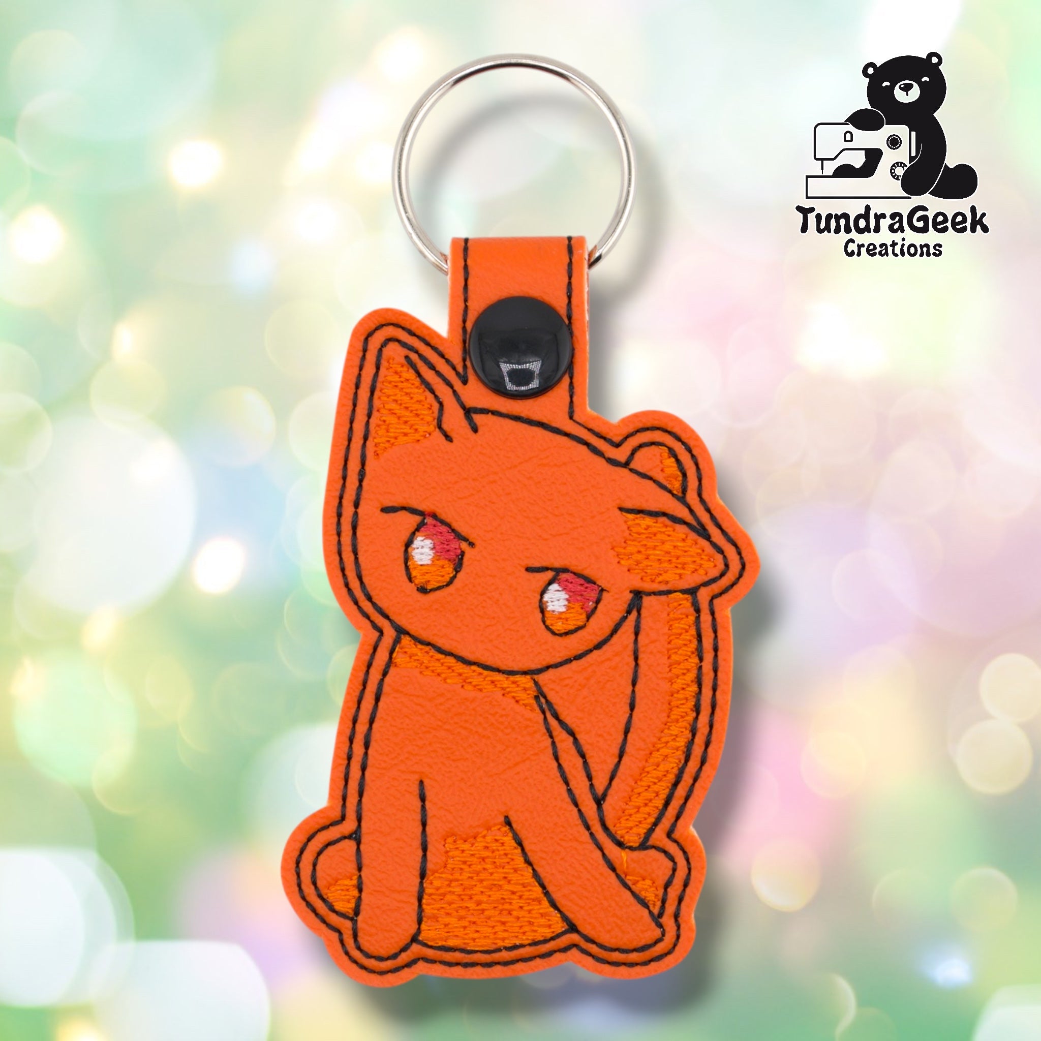 Orange faux leather keychain shaped like a sitting cat with a distinctly unimpressed expression. Black embroidery outlines the body and facial features; eyes are embroidered in orange and white with pink-red detailing. Black snap closure and silver key ring at top. TundraGeek Creations logo in upper right corner.