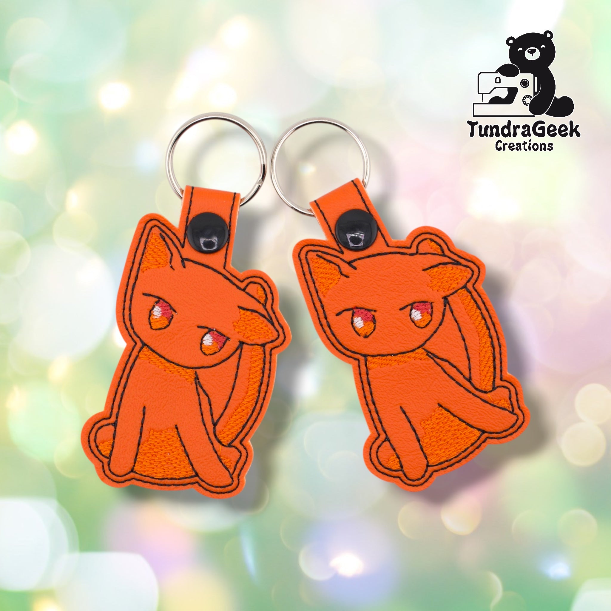 Two orange cat keychains side by side on a pastel bokeh background, showing front and slight three-quarter views of the same design.