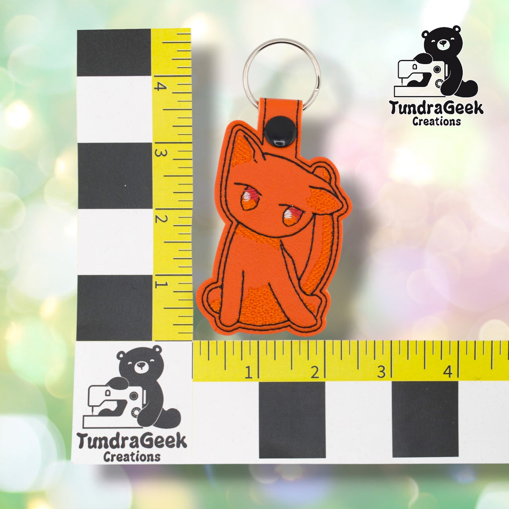 Orange cat keychain positioned against a TundraGeek Creations branded ruler card showing yellow tape measure. Keychain body measures approximately 2.25 inches wide by 3.25 inches tall, not including hardware