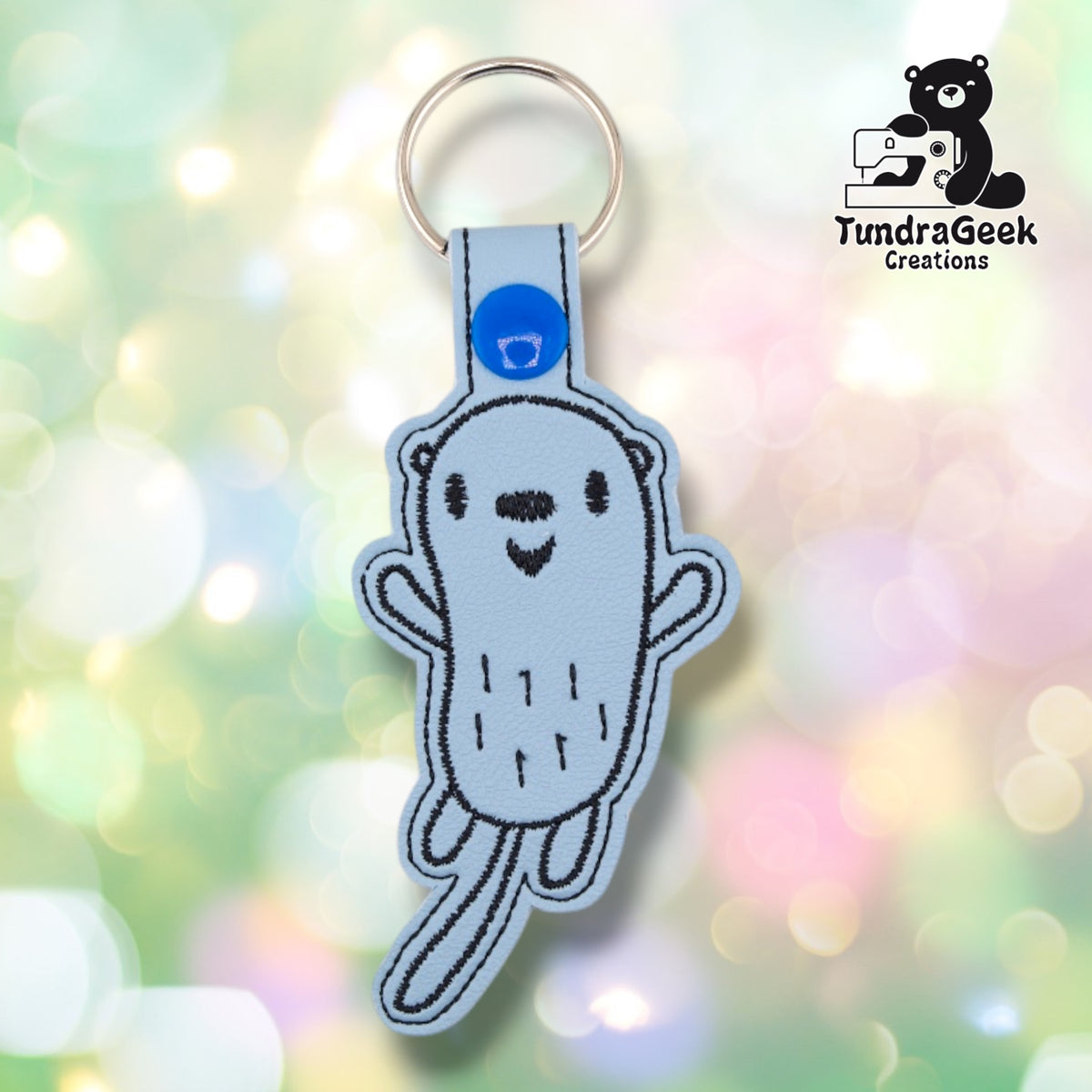 Front of a light blue vinyl keychain embroidered with a simple line-art otter, smiling face, small ears, and paws held up at its sides. Blue plastic snap closure and metal split ring at top.