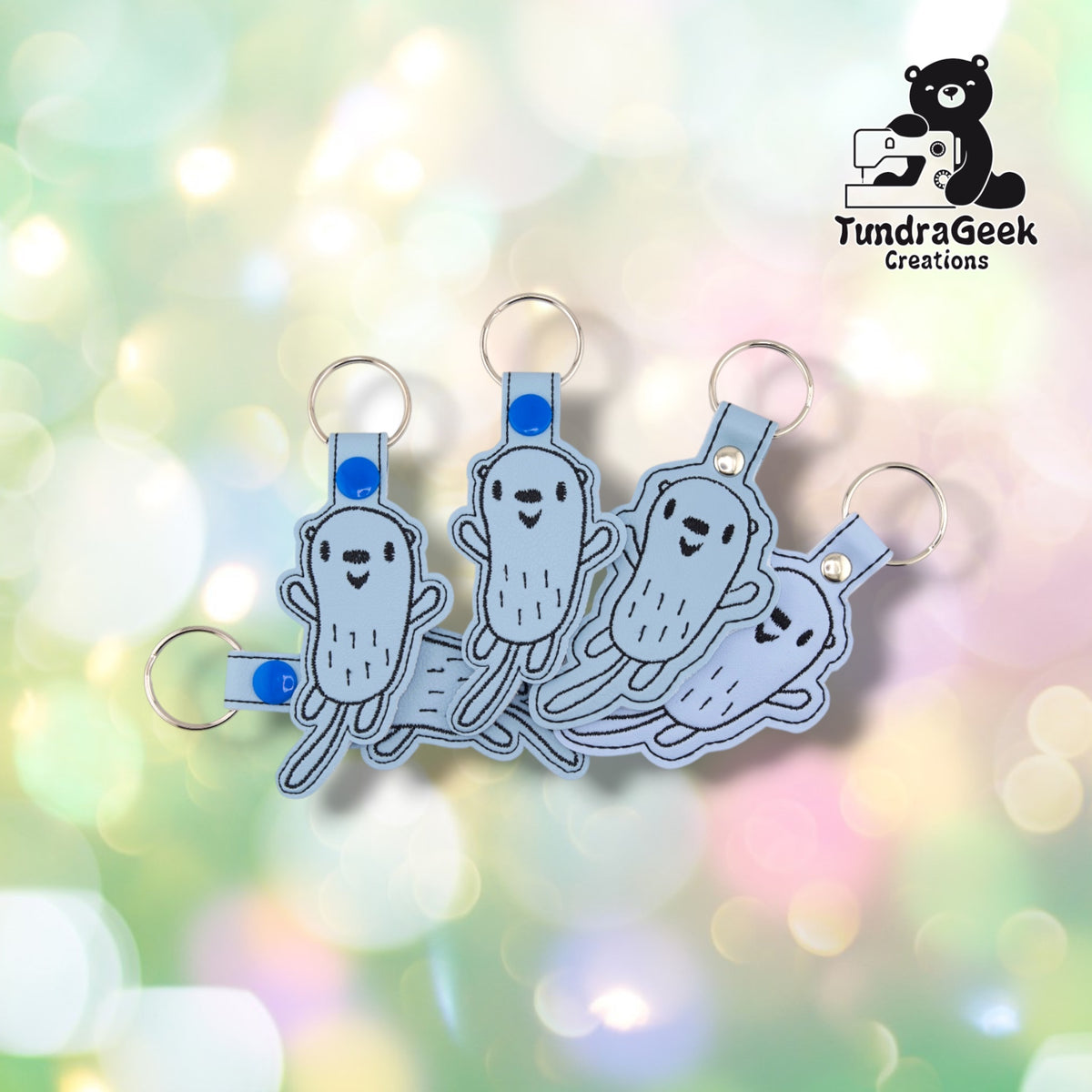 Four identical otter keychains fanned out together, each showing the same light blue vinyl body and line-art otter face and paws.