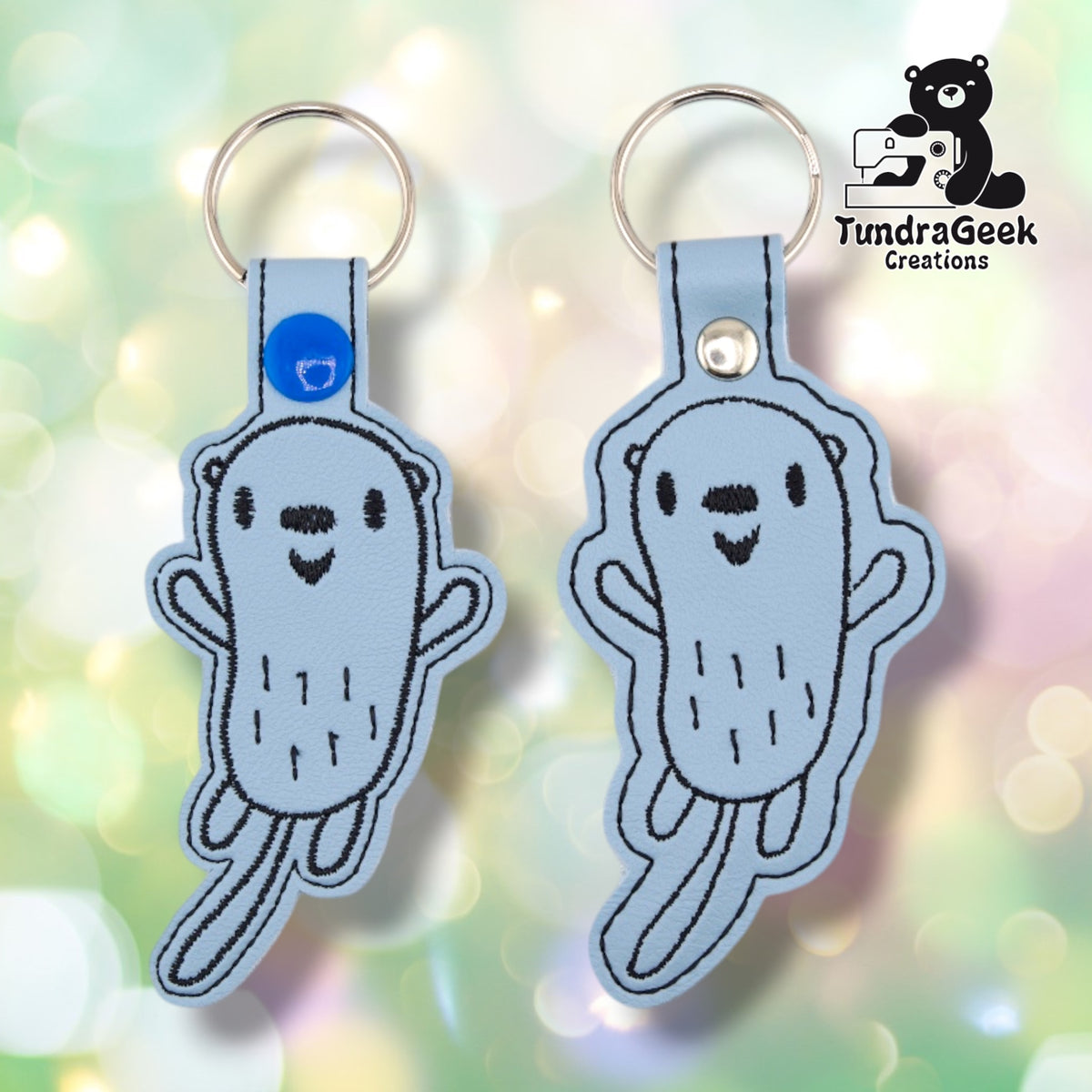 Two otter keychains shown side by side for comparison — the one on the left has a blue plastic snap closure (current production), and the one on the right has a plain silver metal rivet in the same spot (older stock). Both are otherwise identical.
