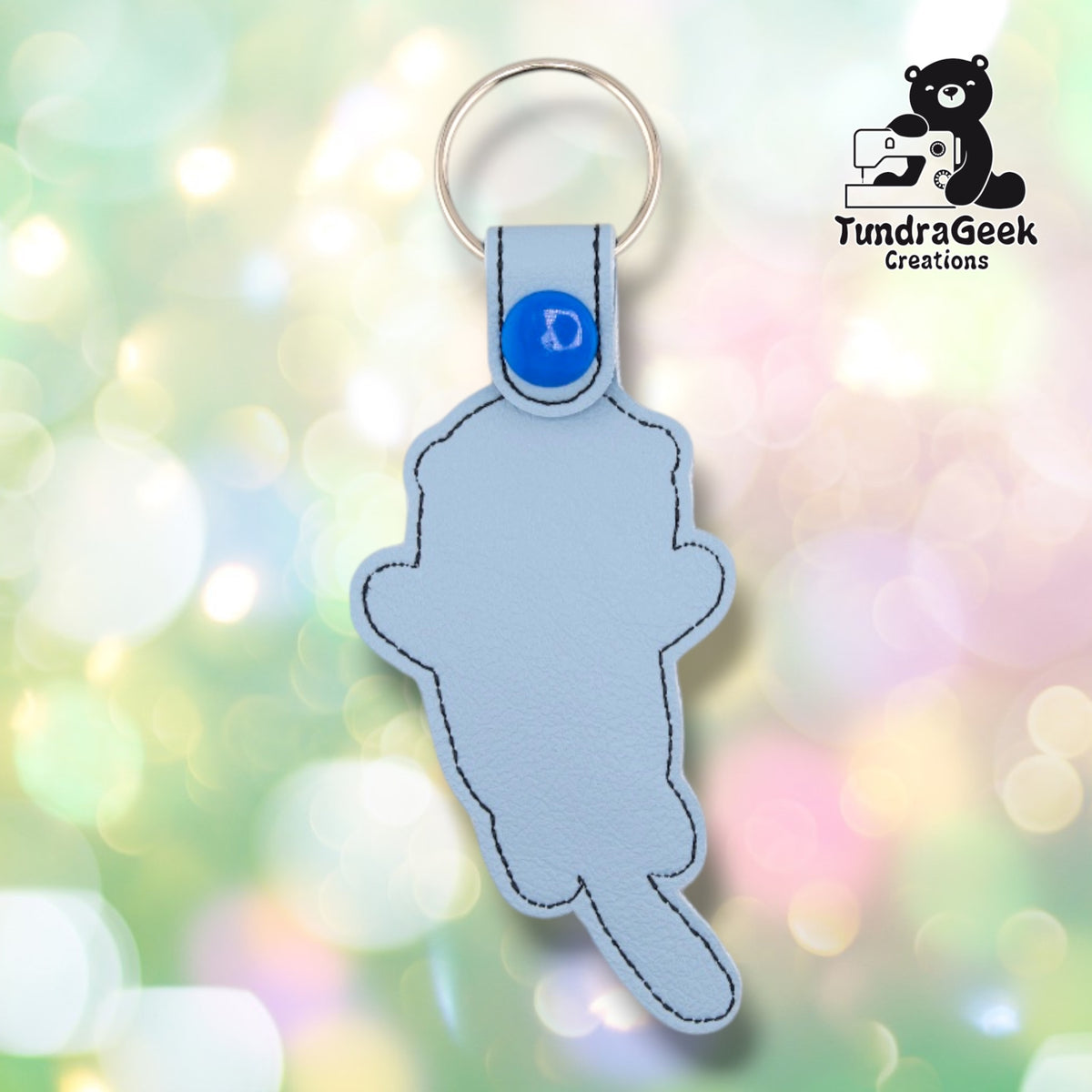 Back of the keychain, showing a plain light blue vinyl reverse side with black outline stitching following the otter's silhouette. Blue plastic snap closure and metal split ring at top.