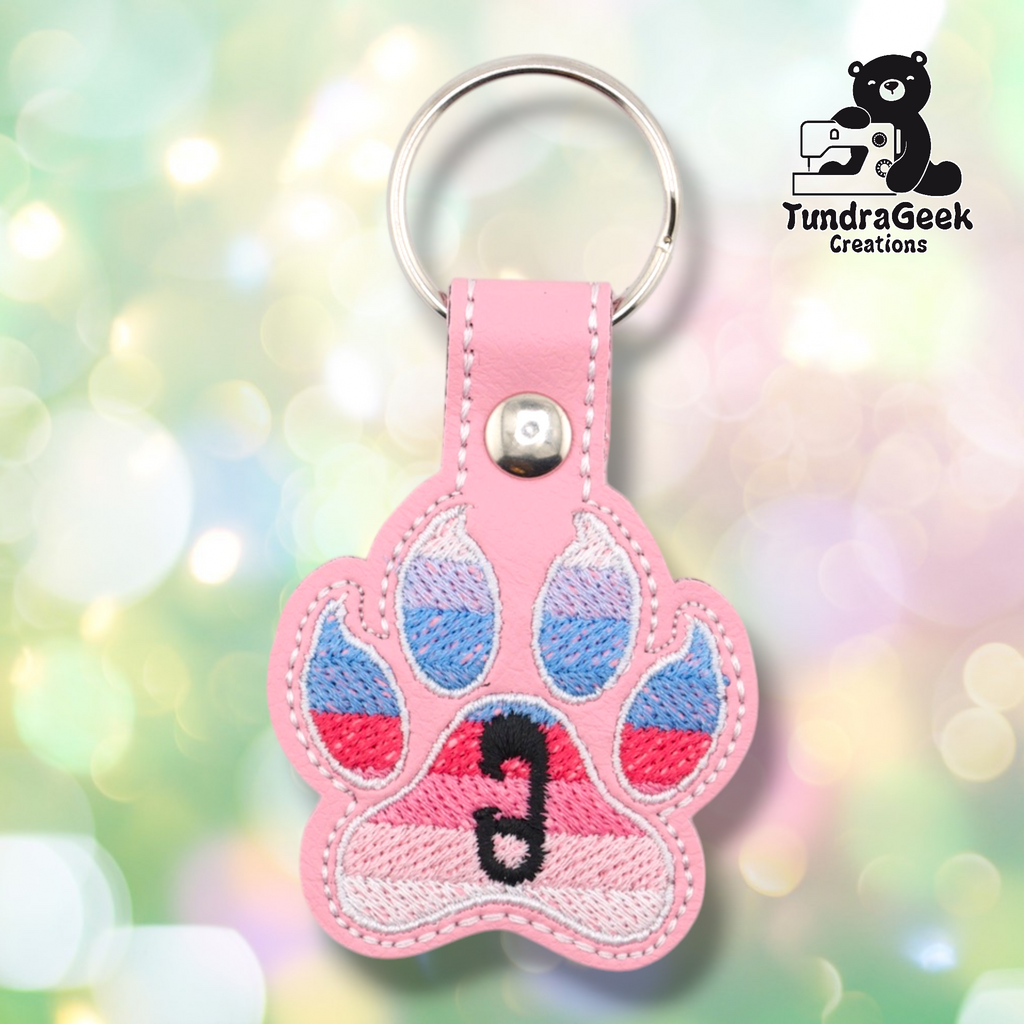 Pink embroidered vinyl paw-shaped keychain featuring the ABDL pride flag colors — horizontal stripes of light blue, periwinkle, red/pink, and soft pink. A small black safety pin is embroidered in the center pad area. Pink vinyl snap tab and silver split ring hardware at top.