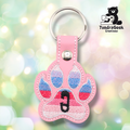Pink embroidered vinyl paw-shaped keychain featuring the ABDL pride flag colors — horizontal stripes of light blue, periwinkle, red/pink, and soft pink. A small black safety pin is embroidered in the center pad area. Pink vinyl snap tab and silver split ring hardware at top.