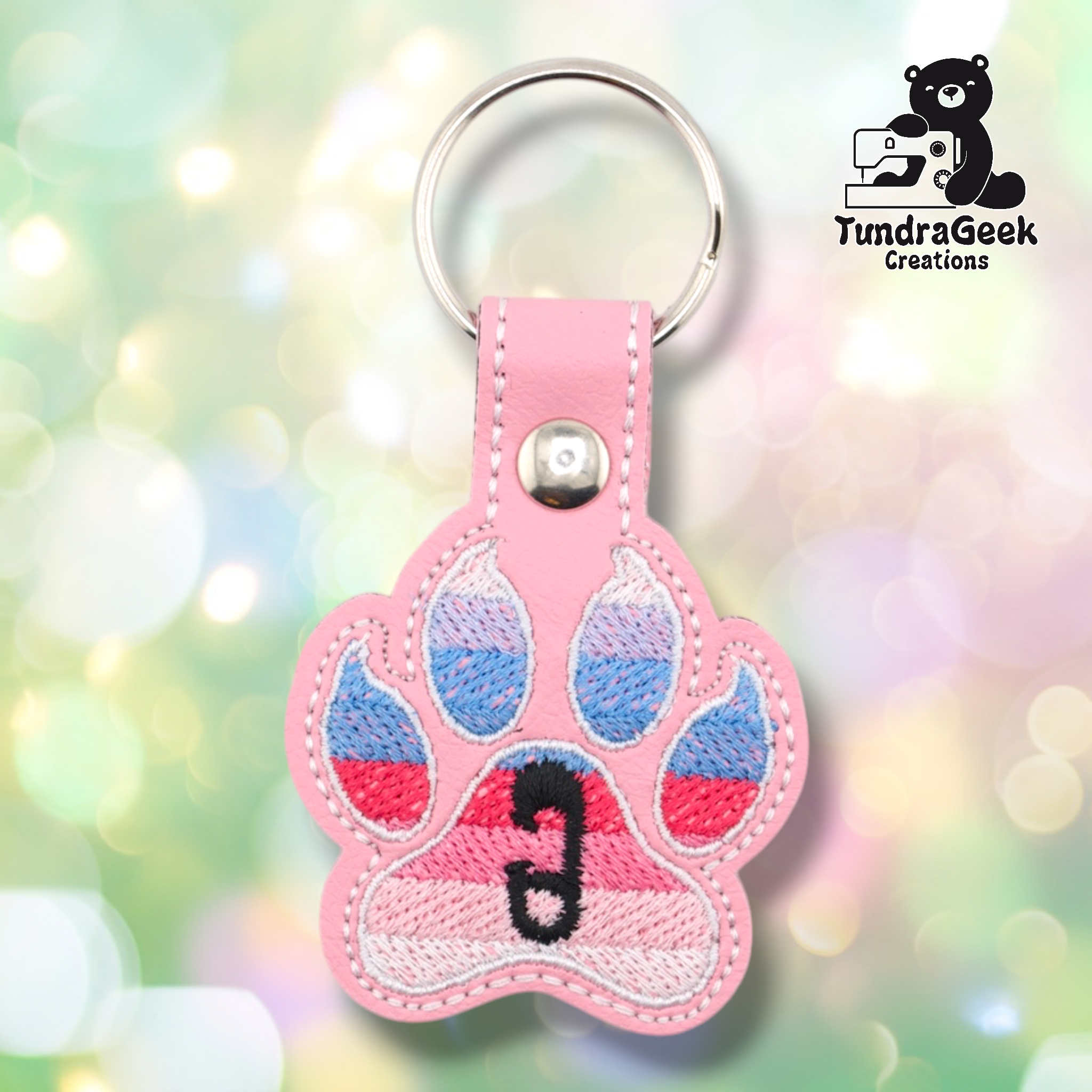 Pink embroidered vinyl paw-shaped keychain featuring the ABDL pride flag colors — horizontal stripes of light blue, periwinkle, red/pink, and soft pink. A small black safety pin is embroidered in the center pad area. Pink vinyl snap tab and silver split ring hardware at top.