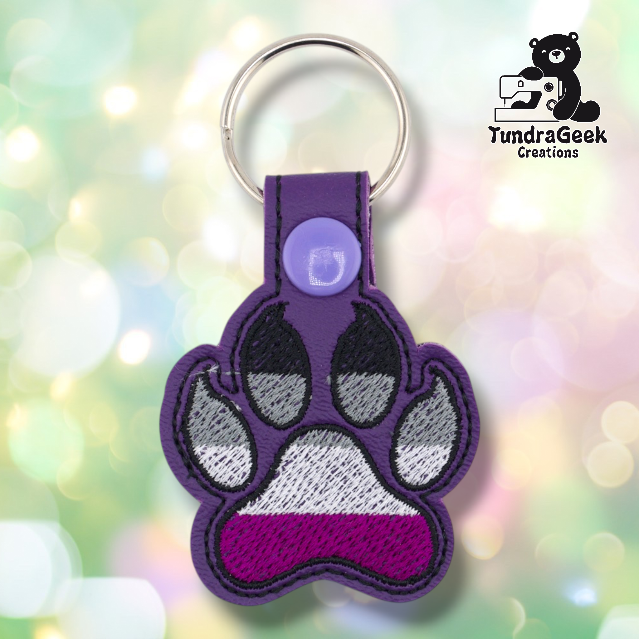 Purple embroidered vinyl paw-shaped keychain featuring asexual pride flag colors — horizontal stripes of black, gray, white, and purple. The paw uses a canine-style design with dark toe bean details outlined in black. Purple vinyl snap tab and silver split ring hardware at top