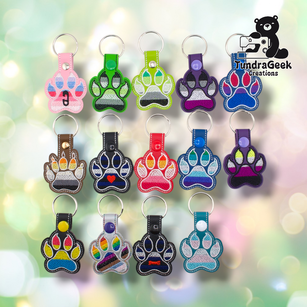 Fourteen embroidered vinyl paw-shaped pride keychains arranged in a three-row grid on a soft bokeh background. The collection includes a wide range of flag colorways. Row one: ABDL (pink), Agender canine-style (dark green), Aromantic (lime green), Asexual (purple), Bisexual (blue). Row two: Bear (brown), Leather (black with heart), Lesbian (red), MLM (blue), Non-Binary (purple). Row three: Pansexual (black), Progress Pride (silver), Pup (black with bone), Transgender (teal). All keychains have silver split 