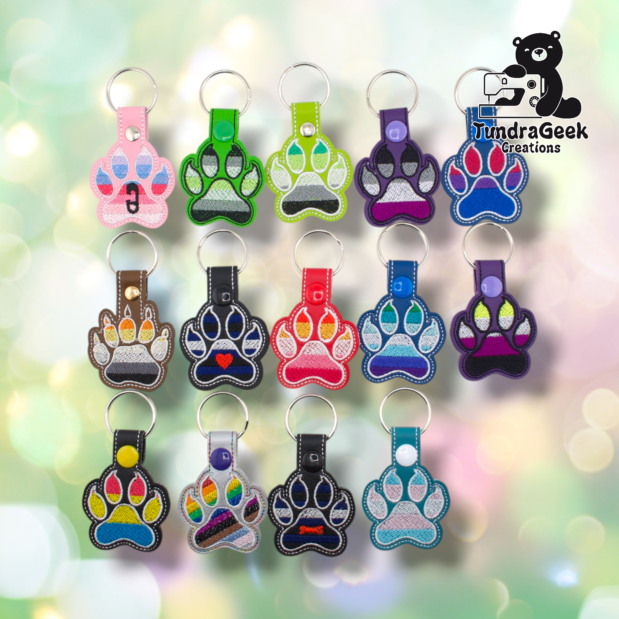 Fourteen embroidered vinyl paw-shaped pride keychains arranged in a three-row grid on a soft bokeh background. The collection includes a wide range of flag colorways. Row one: ABDL (pink), Agender canine-style (dark green), Aromantic (lime green), Asexual (purple), Bisexual (blue). Row two: Bear (brown), Leather (black with heart), Lesbian (red), MLM (blue), Non-Binary (purple). Row three: Pansexual (black), Progress Pride (silver), Pup (black with bone), Transgender (teal). All keychains have silver split 