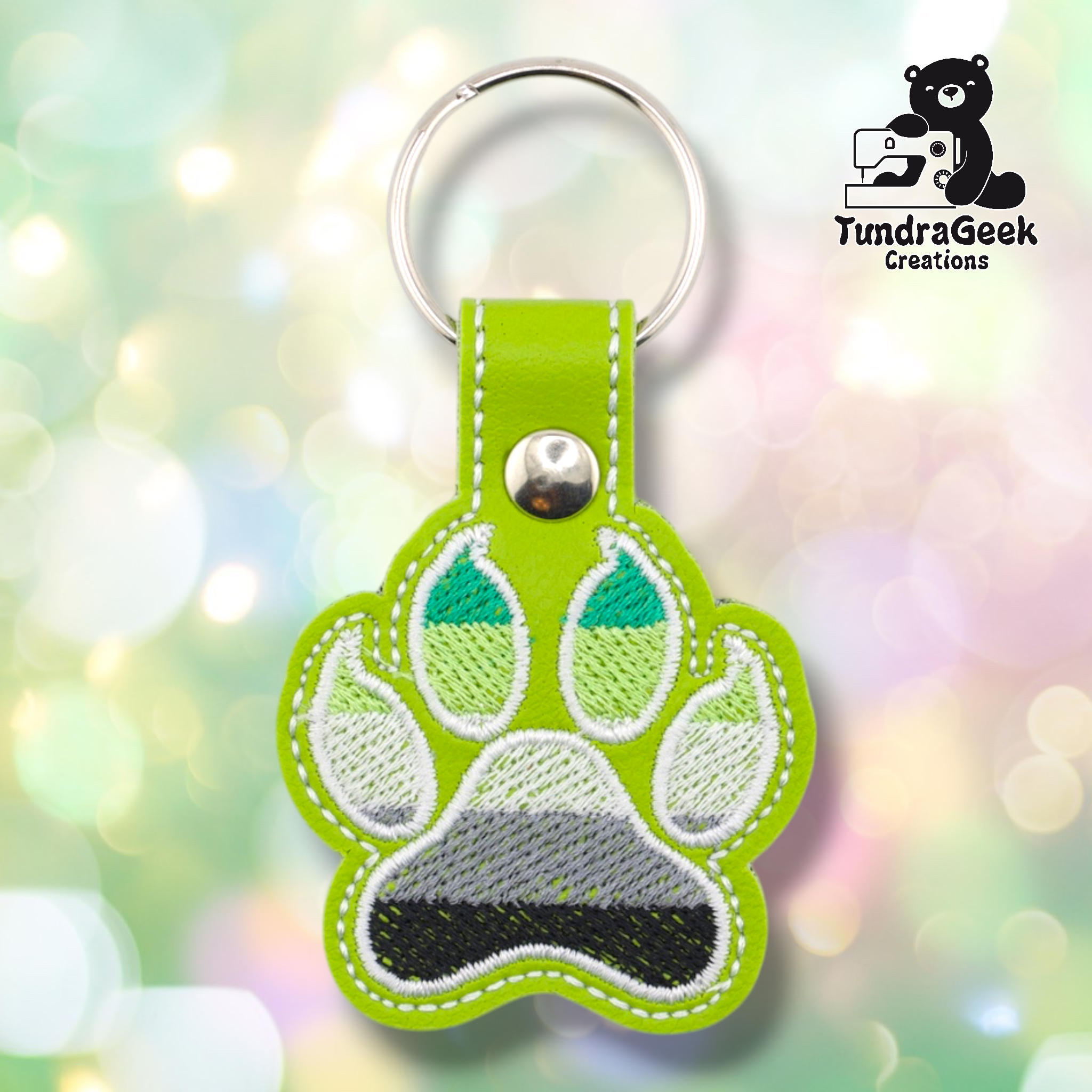 Lime green embroidered vinyl paw-shaped keychain featuring aromantic pride flag colors — horizontal stripes of dark green, light green, white, gray, and black. The paw uses a rounded toe bean design with white outlines. Lime green vinyl snap tab and silver split ring hardware at top.