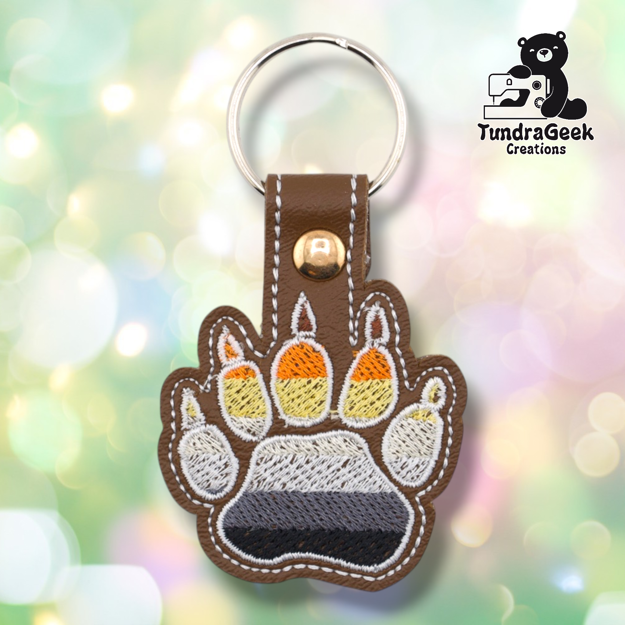 Brown embroidered vinyl paw-shaped keychain featuring bear pride flag colors — horizontal stripes of dark brown, orange, gold/yellow, tan, white, gray, and black. The paw uses a bear claw design with prominent pointed claw tips and white outlines. Brown vinyl snap tab and gold-tone snap hardware at top, silver split ring.
