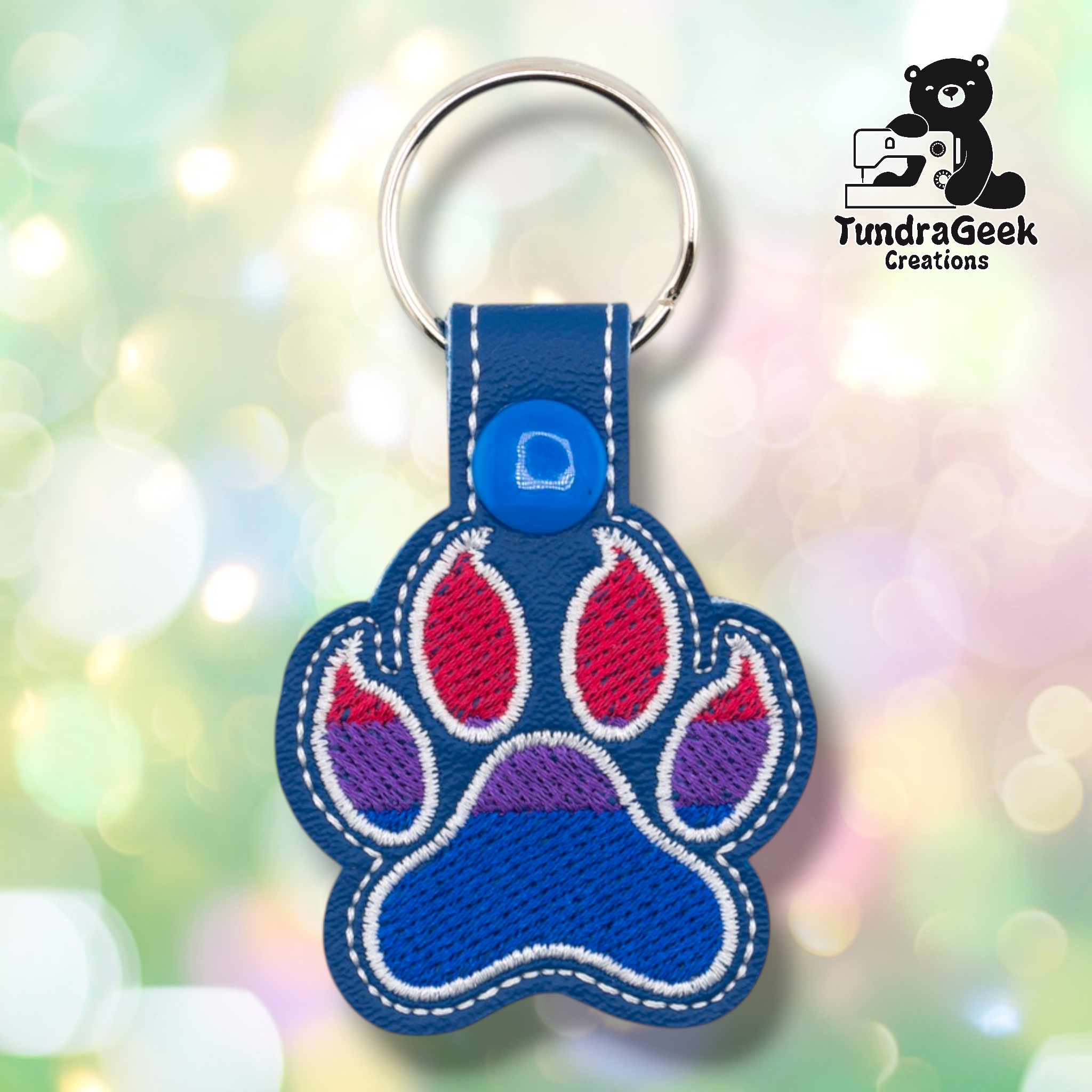 Royal blue embroidered vinyl paw-shaped keychain featuring bisexual pride flag colors — horizontal stripes of pink/magenta, purple, and blue. The paw uses a rounded toe bean design with white outlines on a blue vinyl background. Blue vinyl snap tab and silver split ring hardware at top.