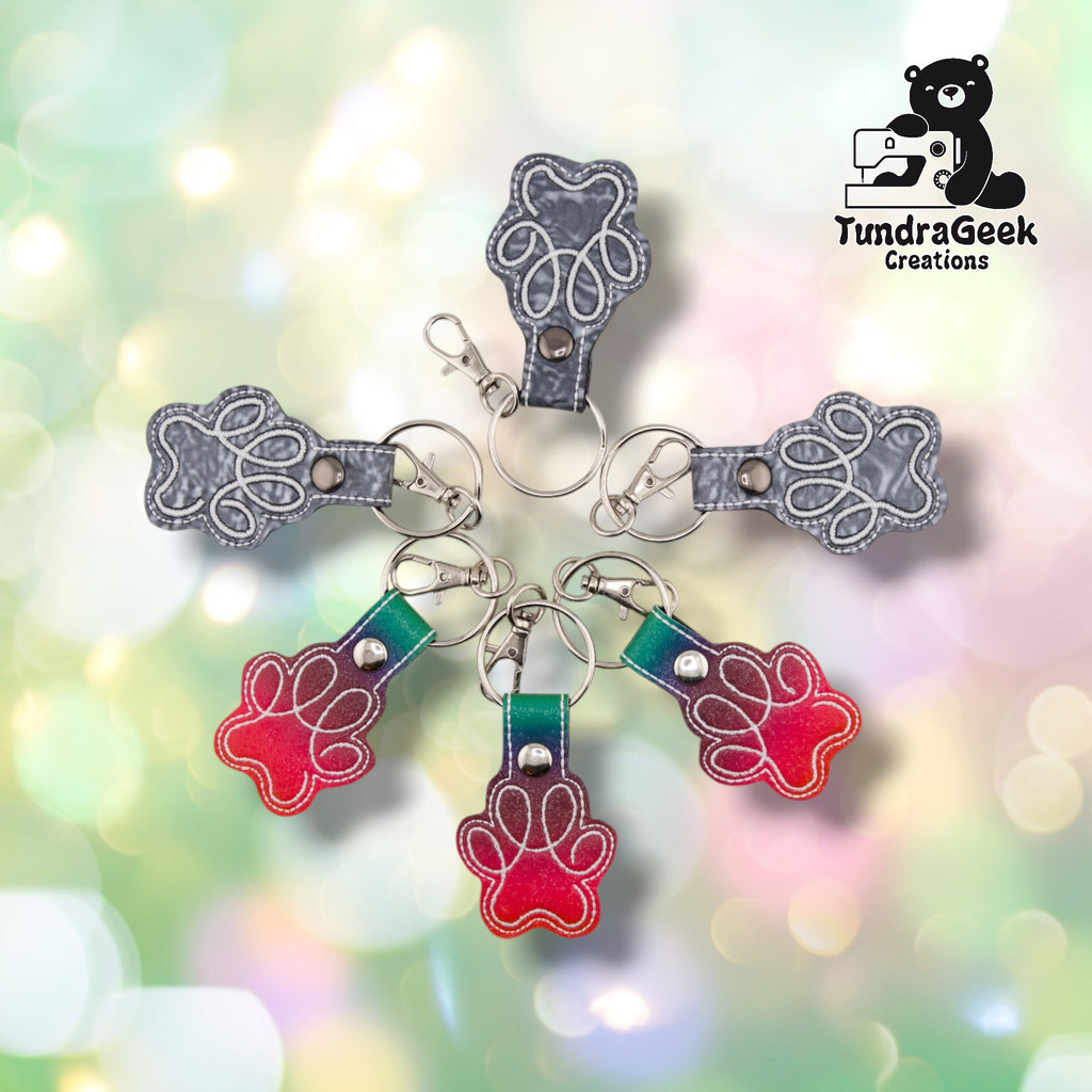 ix paw print keychains arranged in a circle on a soft pastel bokeh background — three in the Silver marbled grey colorway and three in the Rainbow ombre colorway transitioning from teal through purple to red. Each shows the white embroidered paw print stitched outline, split ring, and swivel clip.