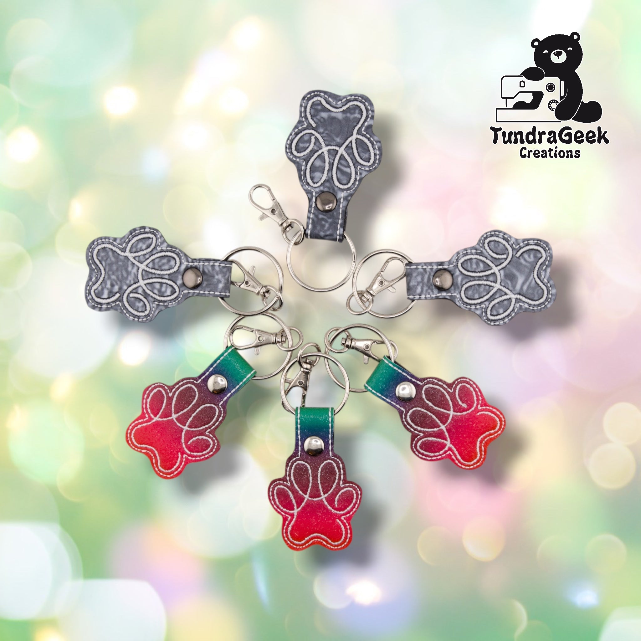 ix paw print keychains arranged in a circle on a soft pastel bokeh background — three in the Silver marbled grey colorway and three in the Rainbow ombre colorway transitioning from teal through purple to red. Each shows the white embroidered paw print stitched outline, split ring, and swivel clip.