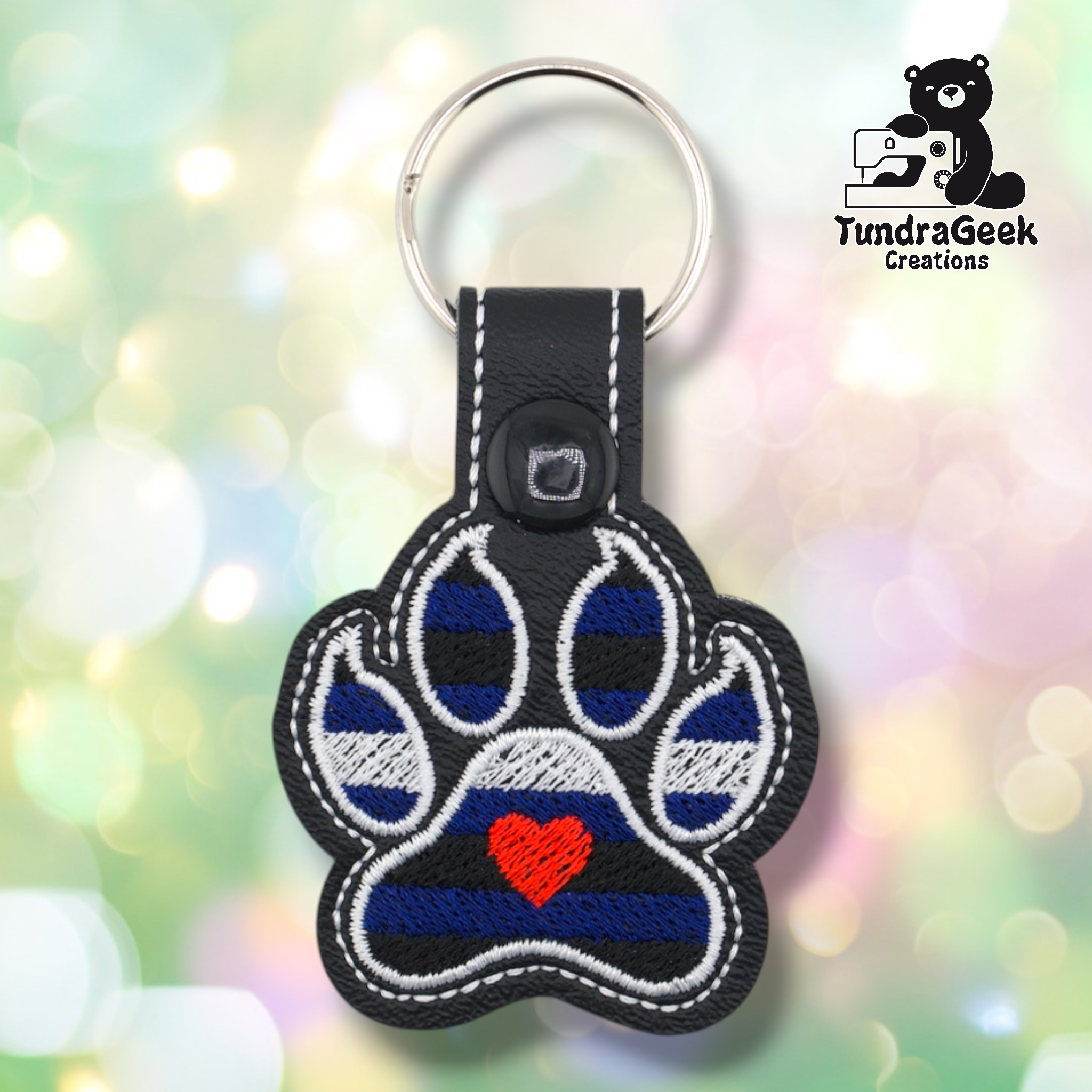 Black embroidered vinyl paw-shaped keychain featuring leather pride flag colors — alternating stripes of royal blue and black with a white stripe across the middle. A small red embroidered heart sits in the center pad area. Black vinyl snap tab and silver split ring hardware at top.