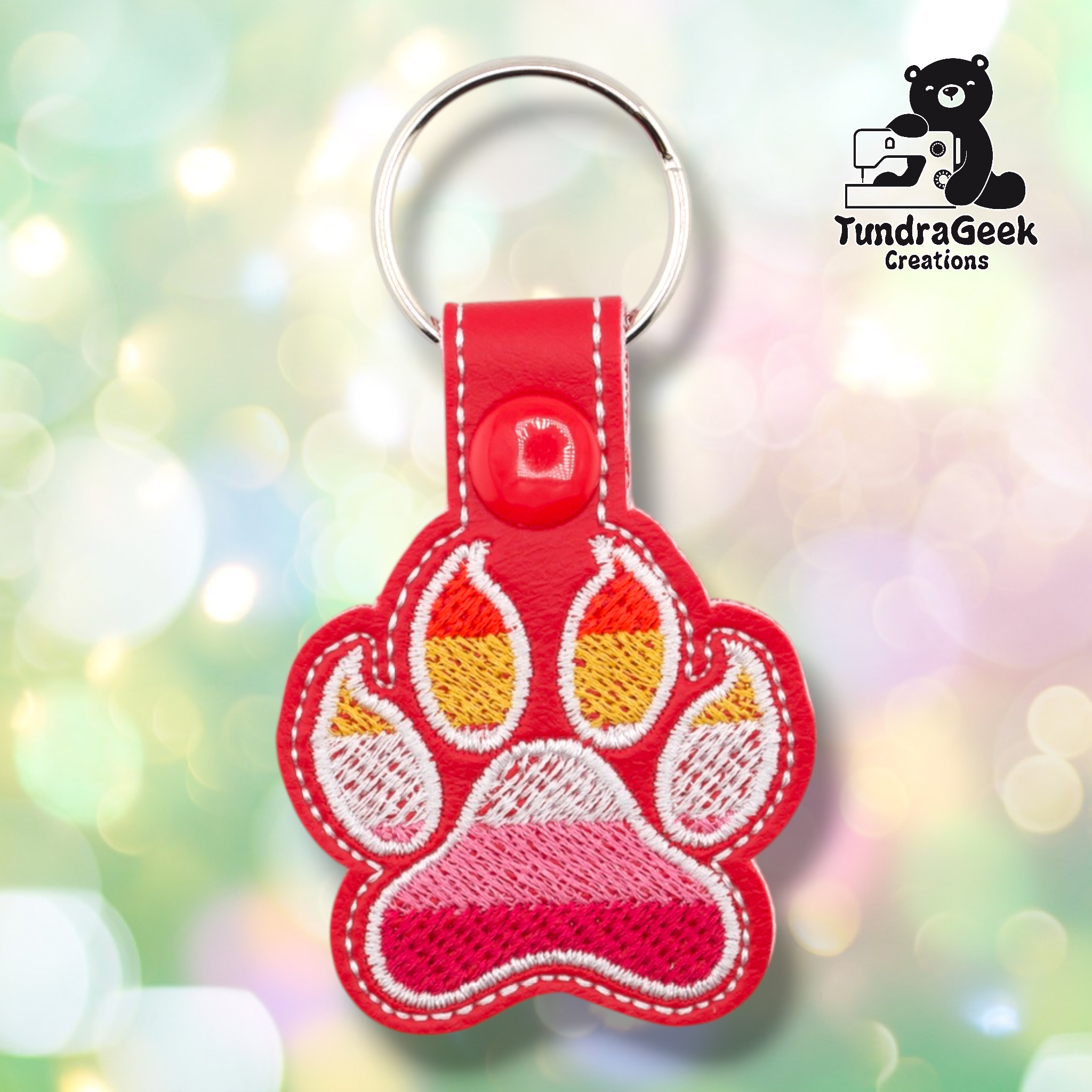 Red embroidered vinyl paw-shaped keychain featuring lesbian pride flag colors — horizontal stripes of dark orange-red, orange, gold, white, pink, and deep pink/red. The paw uses a rounded toe bean design with white outlines on a red vinyl background. Red vinyl snap tab and silver split ring hardware at top