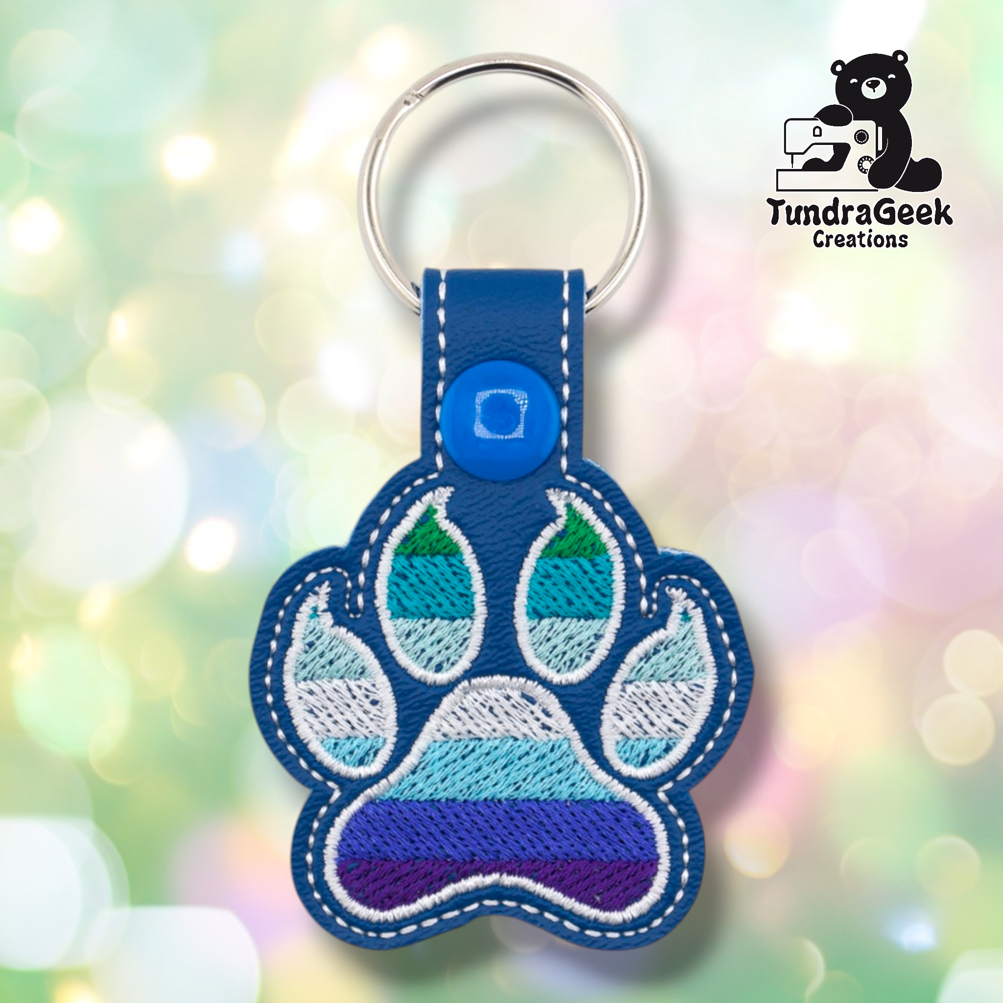 Royal blue embroidered vinyl paw-shaped keychain featuring MLM pride flag colors — horizontal stripes of dark green, teal, light teal/seafoam, white, light blue, periwinkle, and dark indigo/purple. The paw uses a rounded toe bean design with white outlines on a blue vinyl background. Blue vinyl snap tab and blue snap button, silver split ring hardware at top.