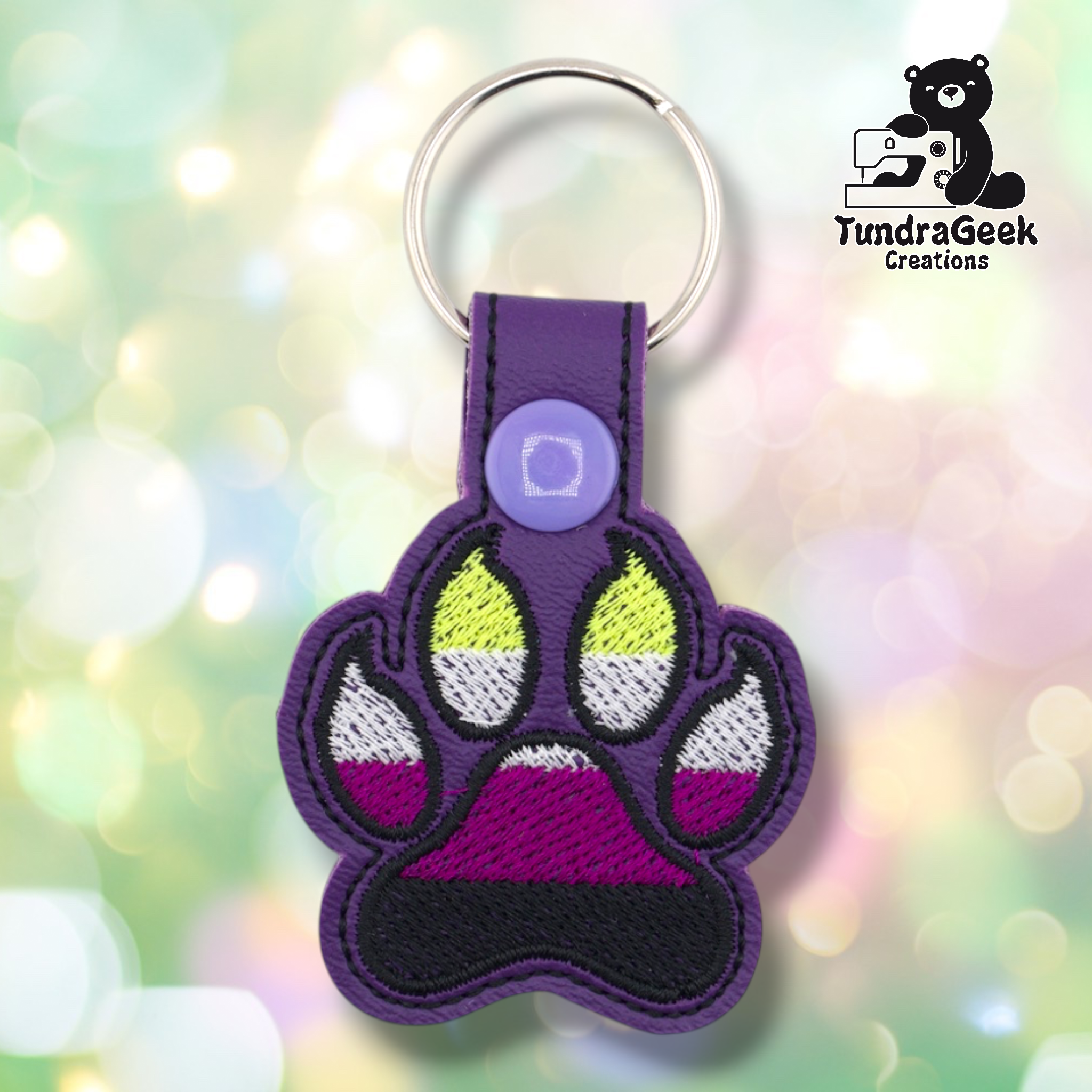 Purple embroidered vinyl paw-shaped keychain featuring non-binary pride flag colors — horizontal stripes of yellow, white, purple, and black. The paw uses a canine-style design with pointed toe details outlined in black. Purple vinyl snap tab and lavender snap button, silver split ring hardware at top.