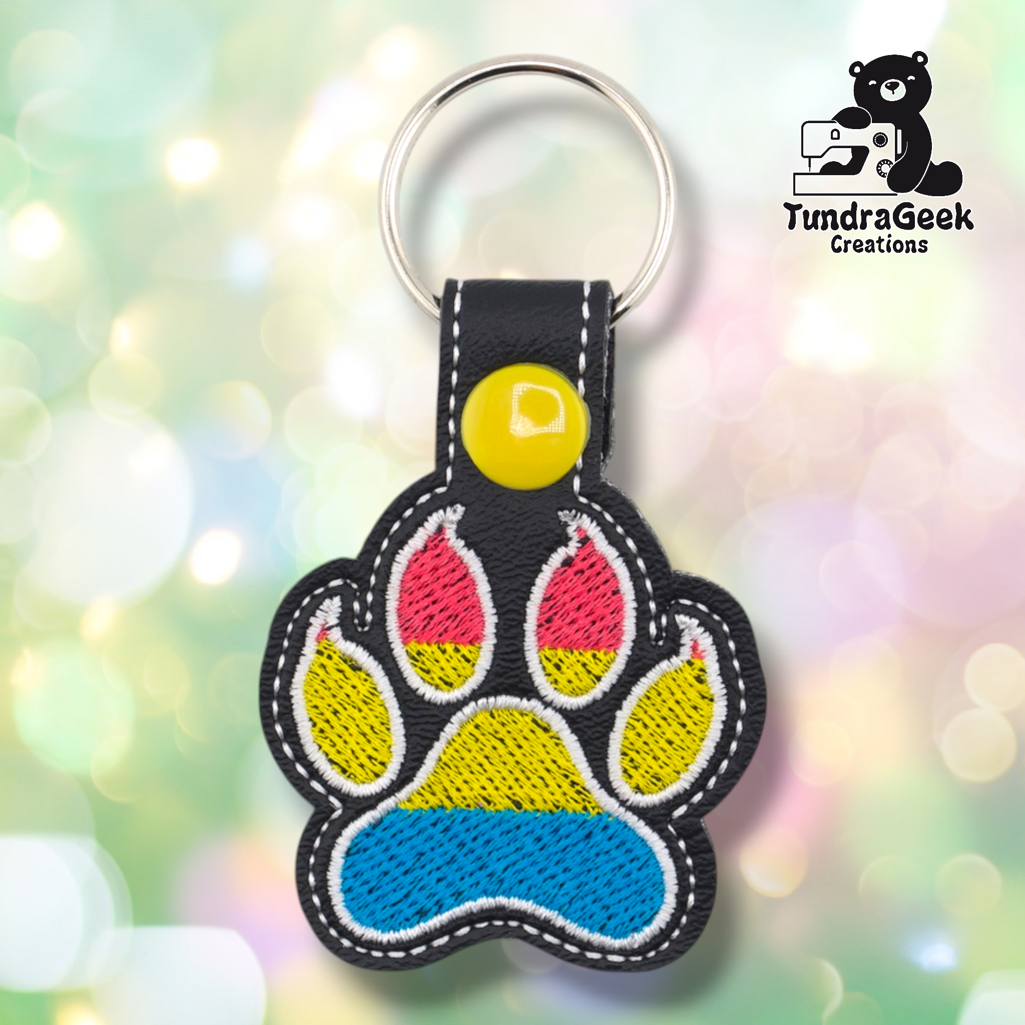 Black embroidered vinyl paw-shaped keychain featuring pansexual pride flag colors — horizontal stripes of hot pink, yellow, and sky blue. The paw uses a canine-style design with pointed toe details outlined in white on a black vinyl background. Black vinyl snap tab and yellow snap button, silver split ring hardware at top.