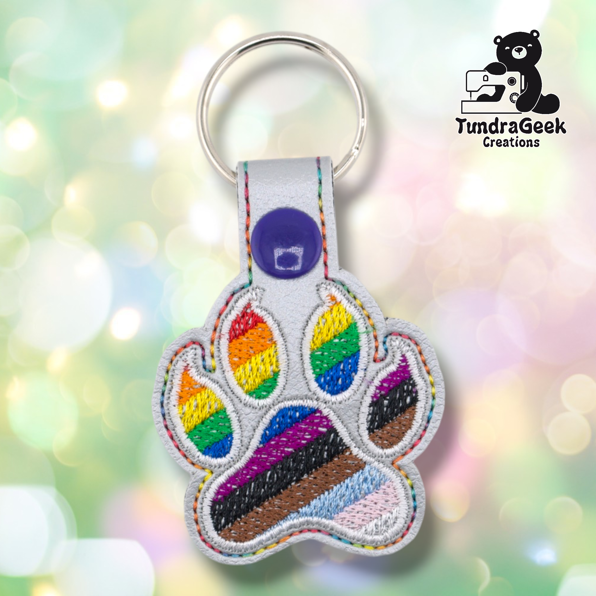Silver/gray embroidered vinyl paw-shaped keychain featuring the Progress Pride flag design — rainbow stripes of red, orange, yellow, green, blue, and purple in the toe beans, with a diagonal chevron of blue, purple, black, brown, light blue, pink, and white across the main pad. Silver vinyl snap tab and purple snap button, silver split ring hardware at top.