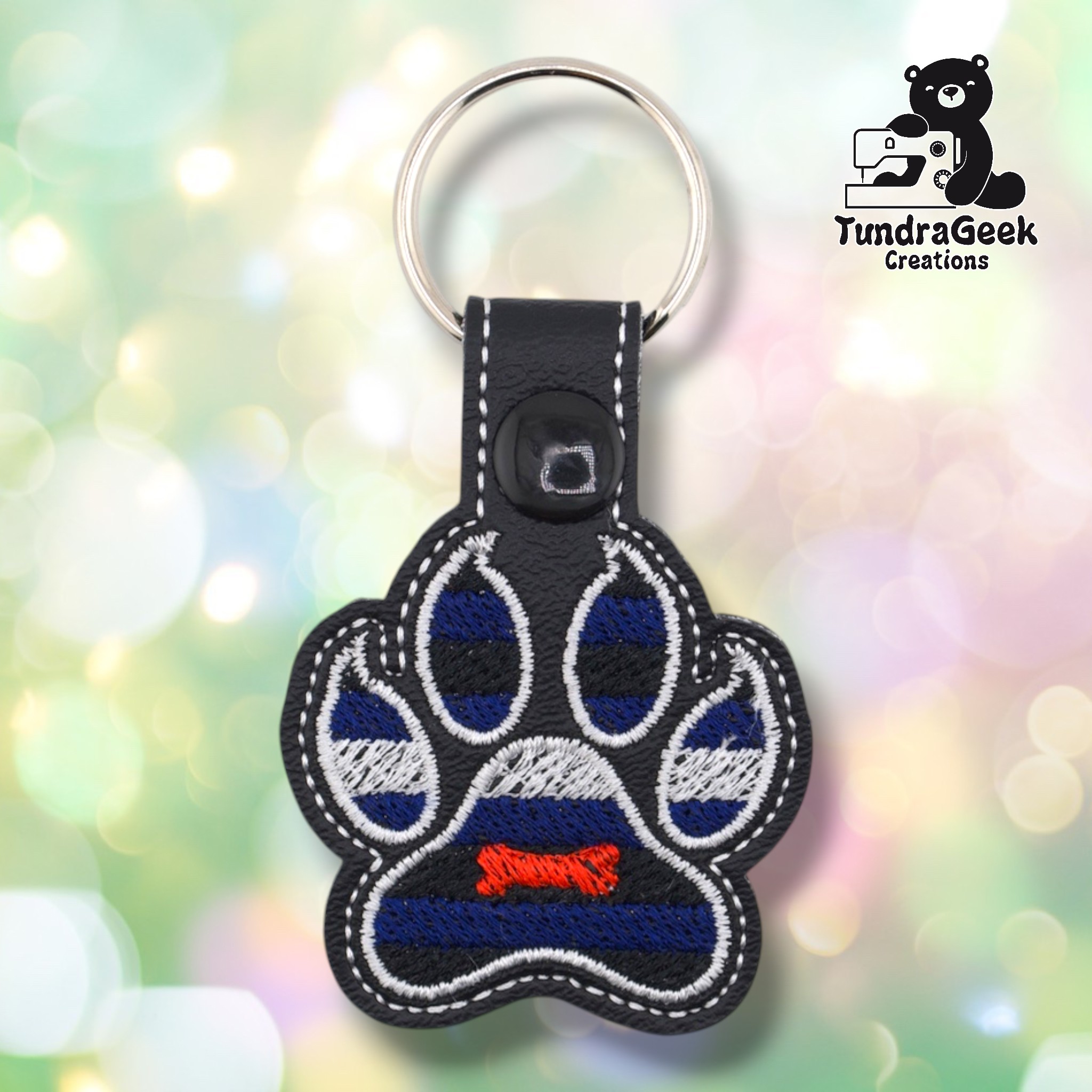 Black embroidered vinyl paw-shaped keychain featuring pup pride flag colors — alternating stripes of royal blue and black with a white stripe across the middle. A small red embroidered bone sits in the center pad area. Black vinyl snap tab and black snap button, silver split ring hardware at top.