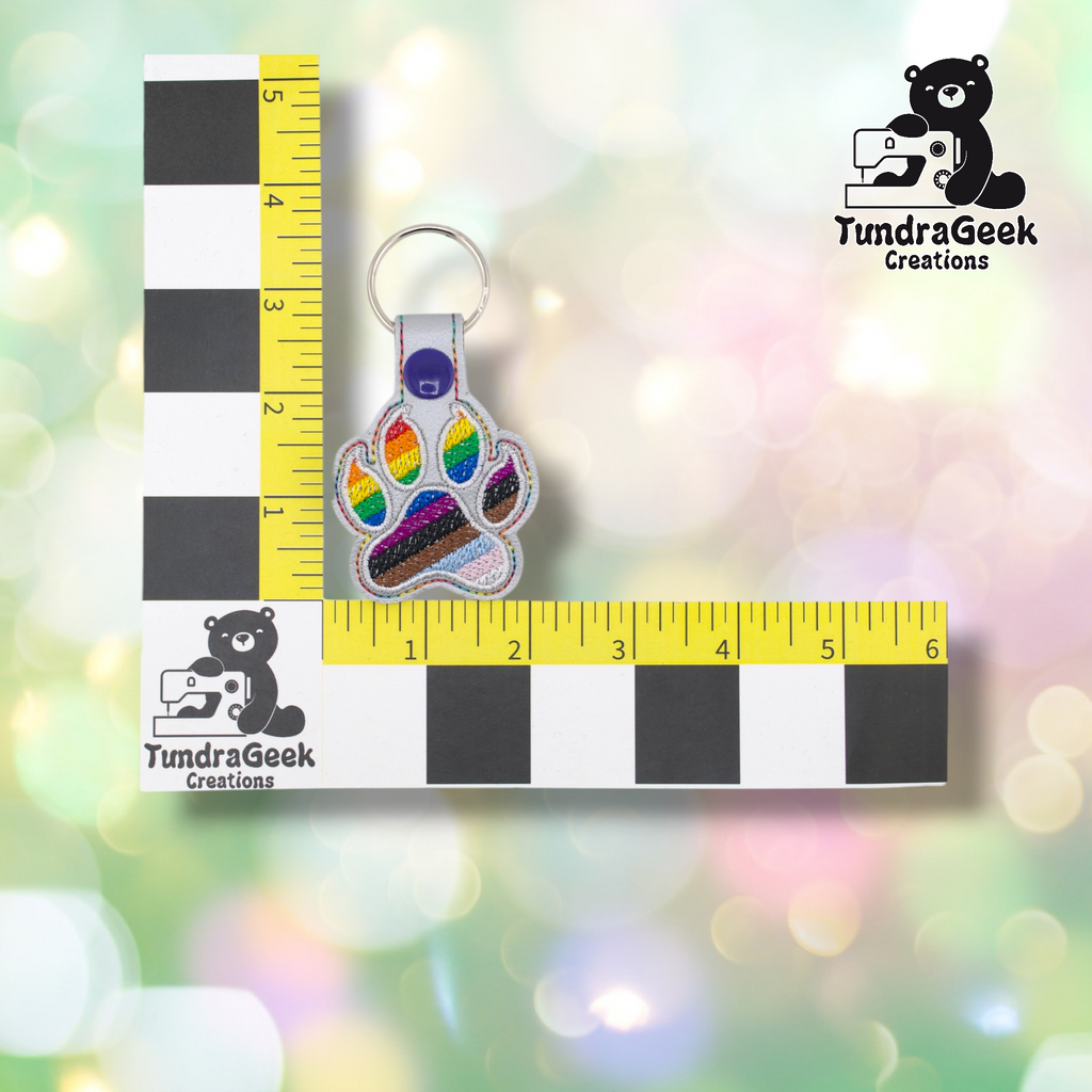 Progress Pride paw keychain shown against a black-and-white checkered background with yellow rulers for scale. The keychain measures approximately 2 inches wide and 3.75 inches tall including hardware. Representative of the size of all variants in this listing.