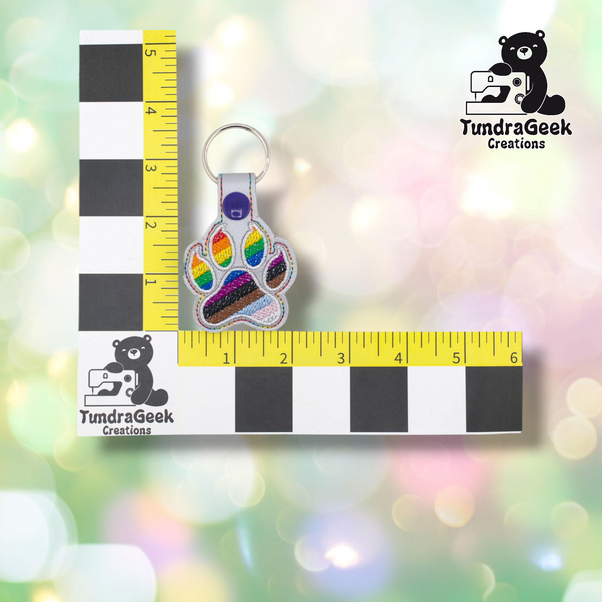 Progress Pride paw keychain shown against a black-and-white checkered background with yellow rulers for scale. The keychain measures approximately 2 inches wide and 3.75 inches tall including hardware. Representative of the size of all variants in this listing.