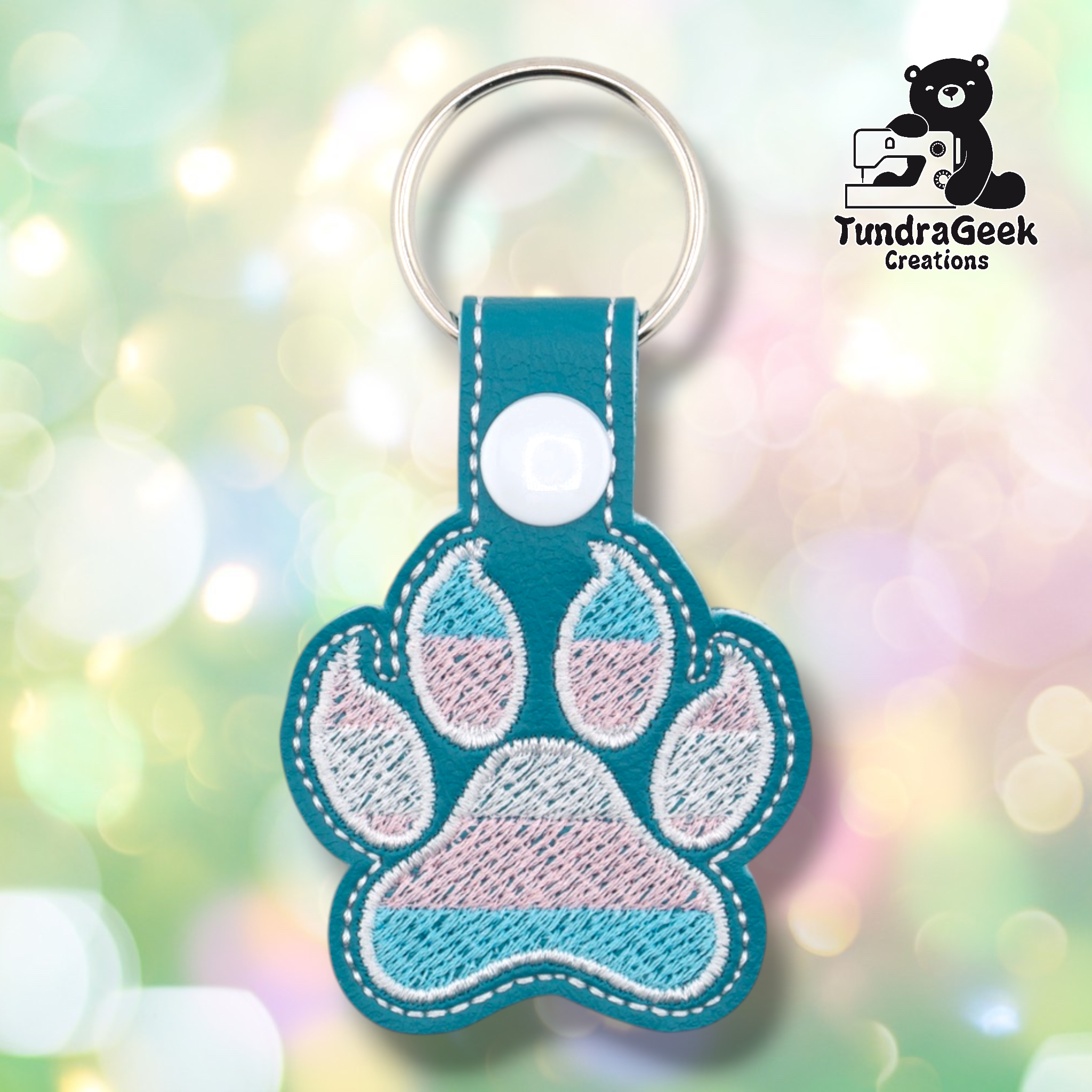 Teal embroidered vinyl paw-shaped keychain featuring transgender pride flag colors — horizontal stripes of light blue, pink, and white. The paw uses a rounded toe bean design with white outlines on a teal vinyl background. Teal vinyl snap tab and white snap button, silver split ring hardware at top