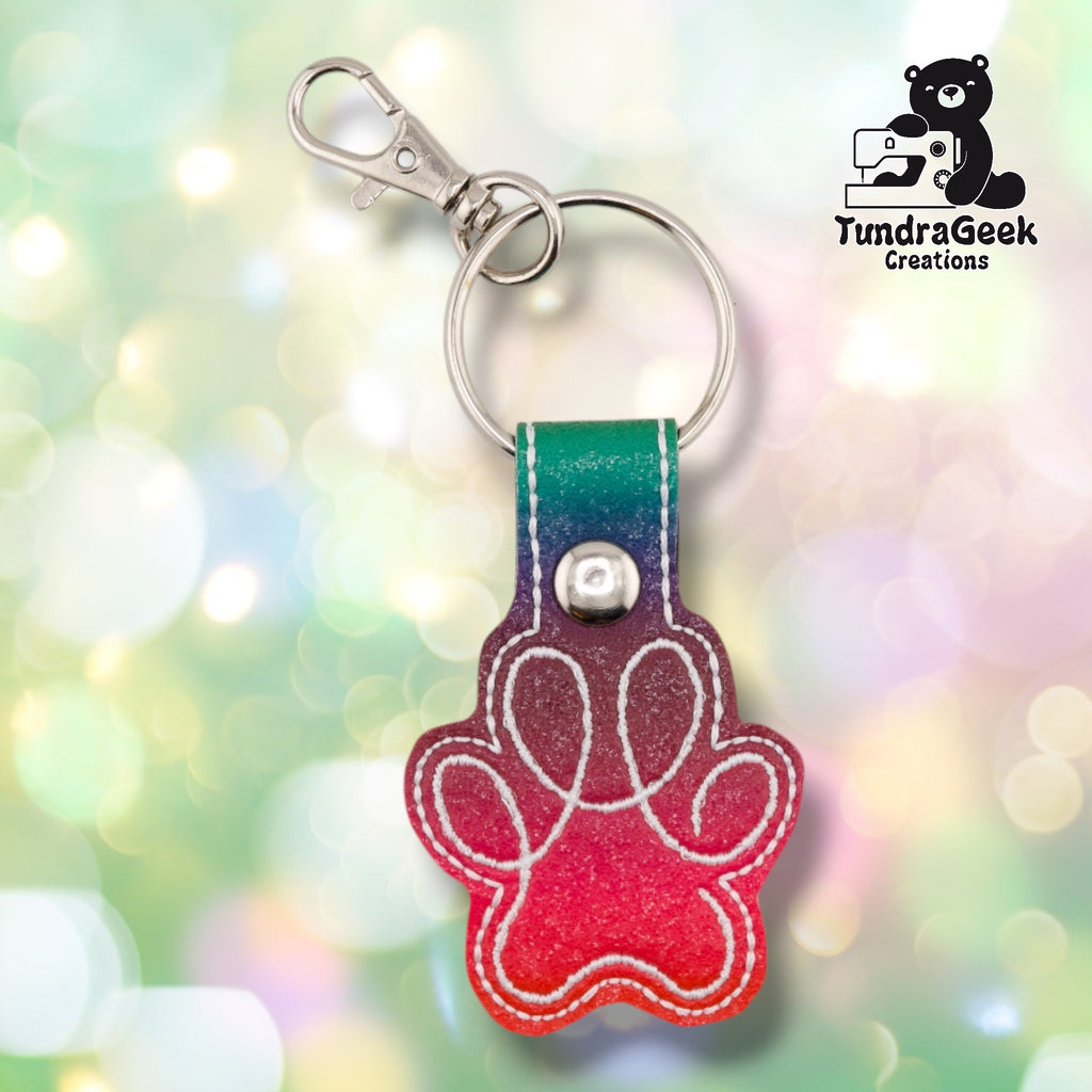 The Rainbow colorway paw print keychain shown flat, with a white embroidered paw print stitched outline over a teal-to-red ombre vinyl charm and a matching teal-to-purple-to-red ombre tab secured with a metal rivet.