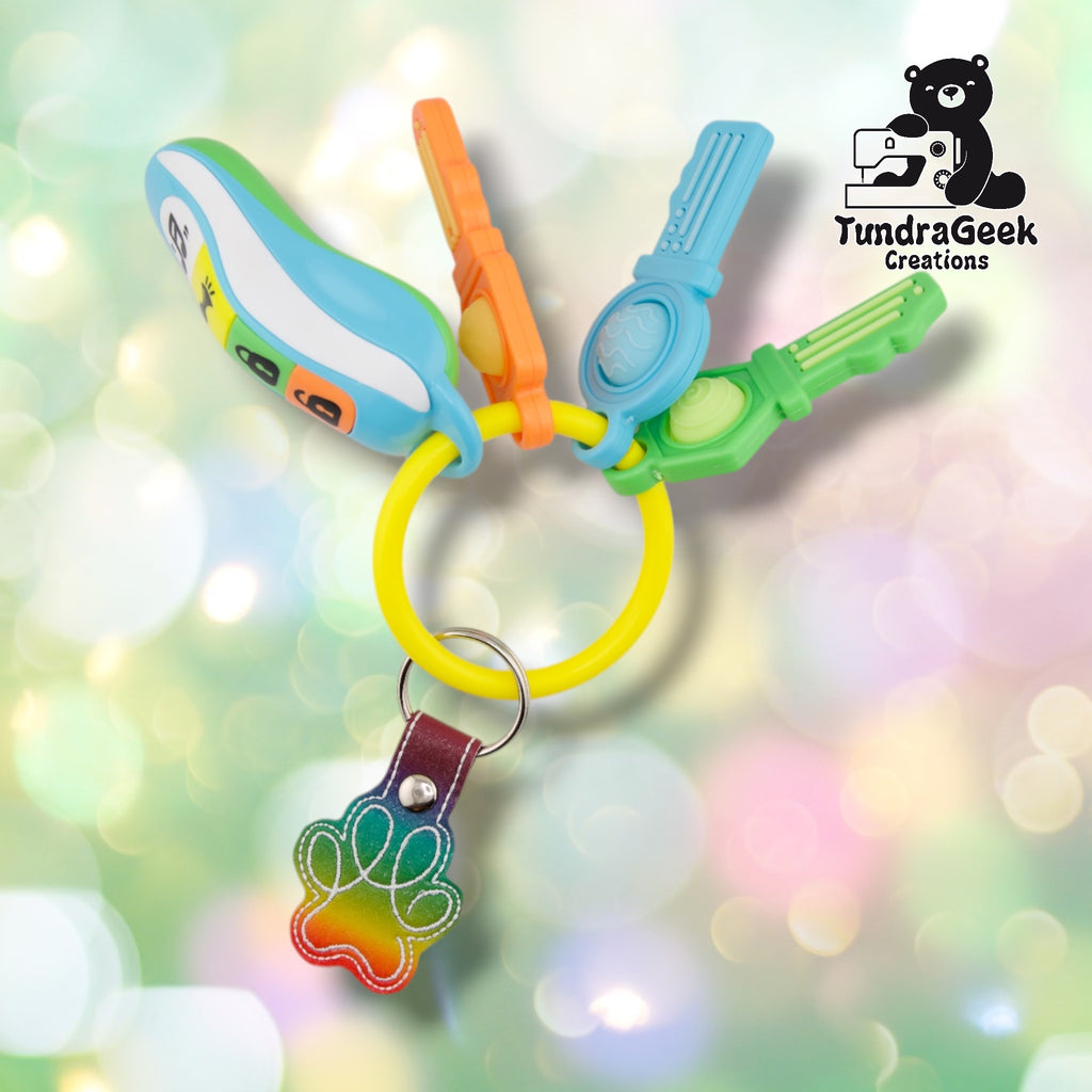 The Rainbow colorway paw print keychain attached via split ring and swivel clip to a colorful set of toy teether keys, showing a green-to-red ombre paw print charm with white stitched outline detailing.