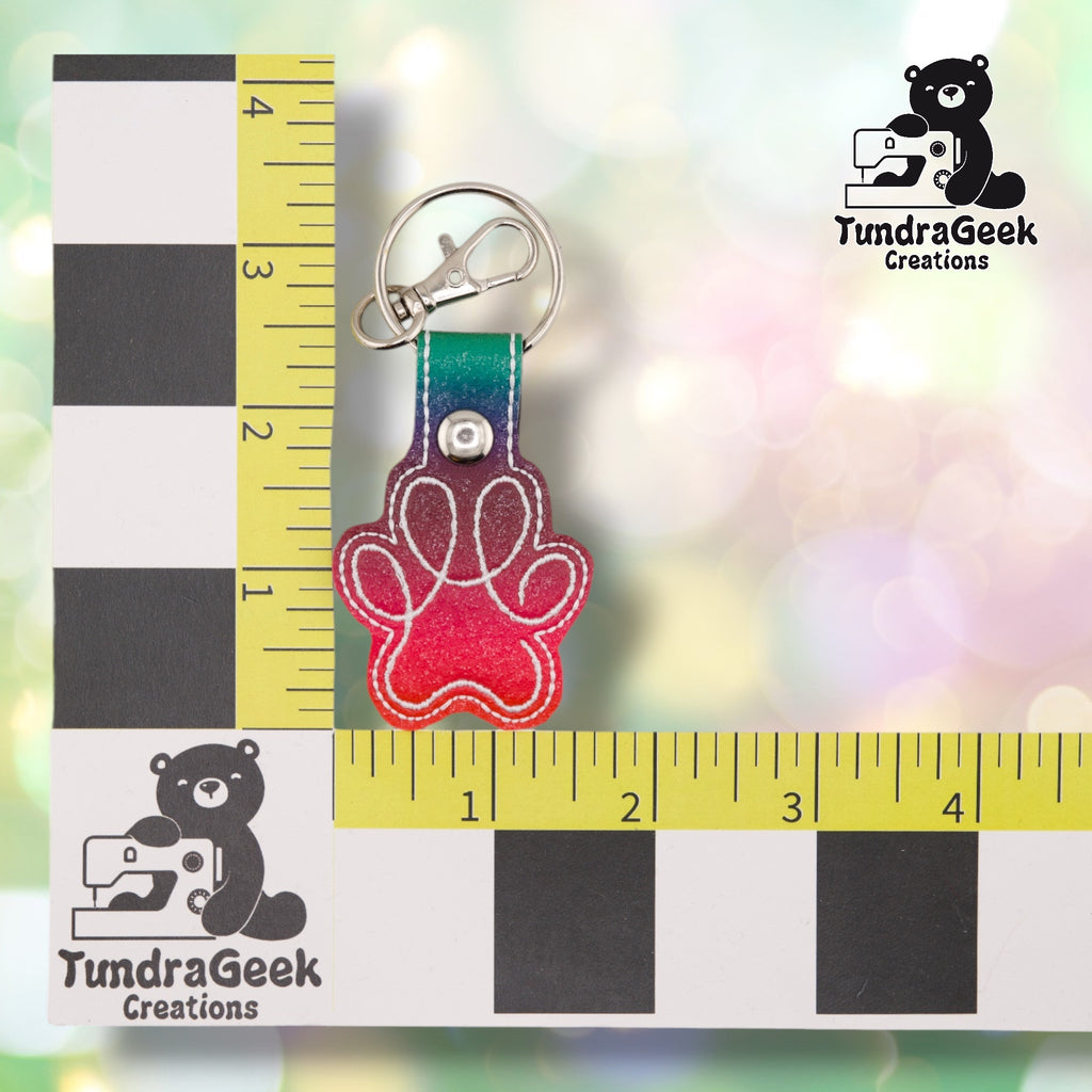 The Rainbow colorway paw print keychain shown against a black-and-white checkered background with yellow rulers for scale, measuring 1.5 inches wide by 3.5 inches tall including hardware. White embroidered paw print outline over teal-to-red ombre vinyl, teal-to-purple-to-red ombre tab, metal rivet closure, and silver split ring with swivel clip.
