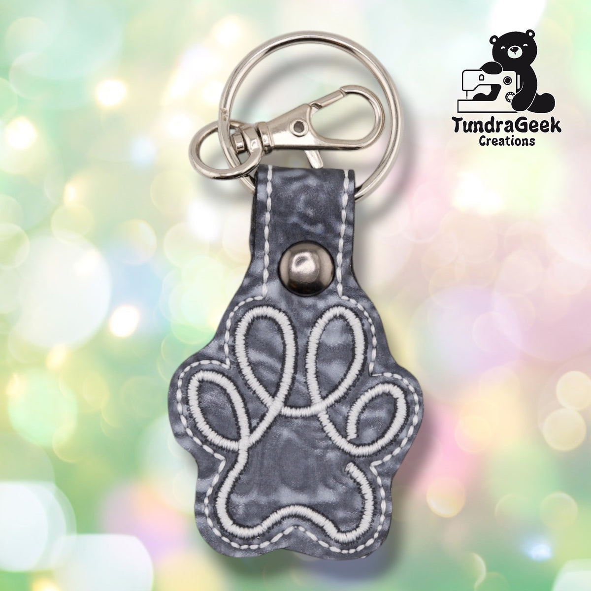 The Silver colorway paw print keychain shown flat, with a white embroidered paw print stitched outline over a marbled grey "Silver Fluidity" vinyl charm and matching grey tab secured with a metal rivet.