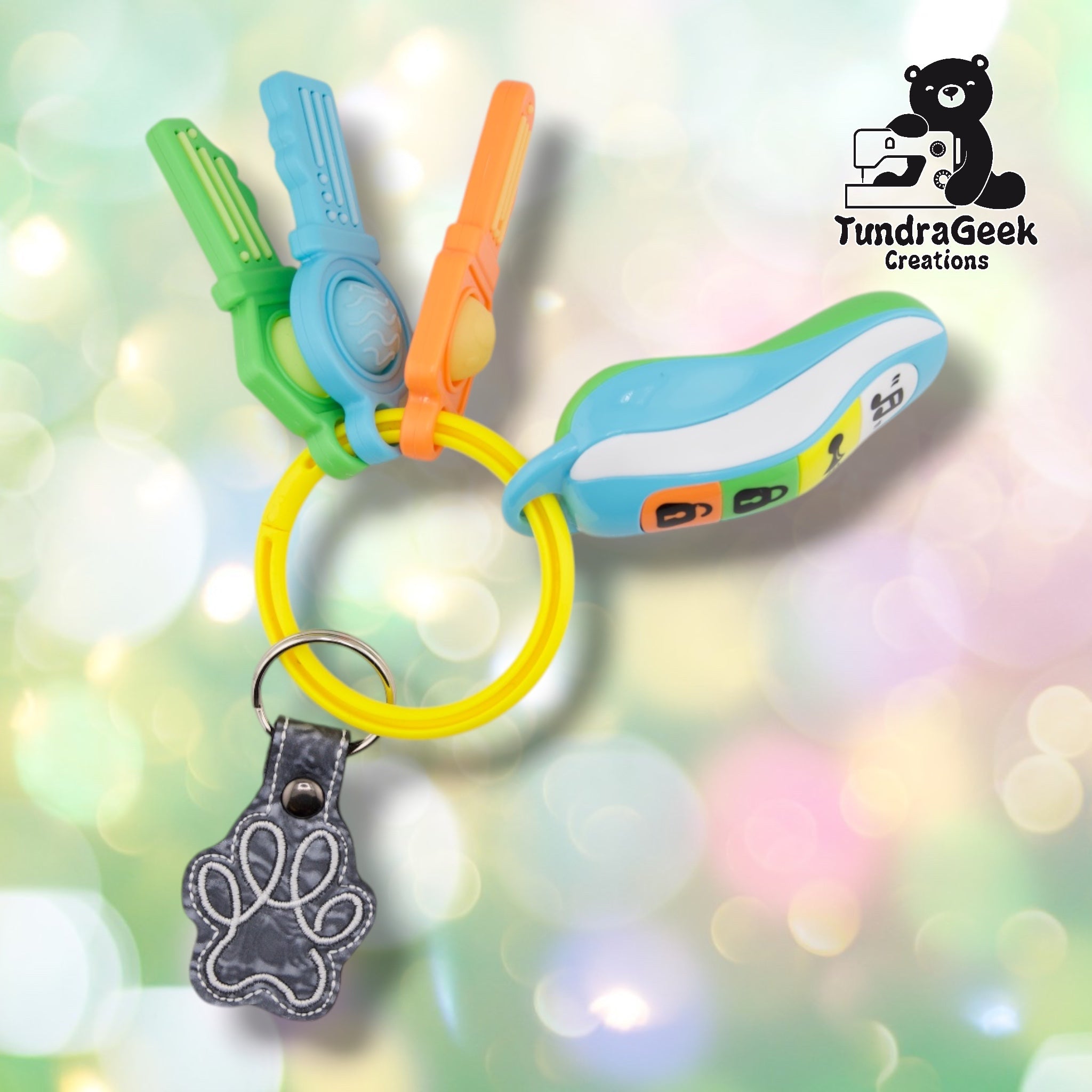 The Silver colorway paw print keychain attached via split ring and swivel clip to a colorful set of toy teether keys, showing a marbled grey paw print charm with white stitched outline detailing.