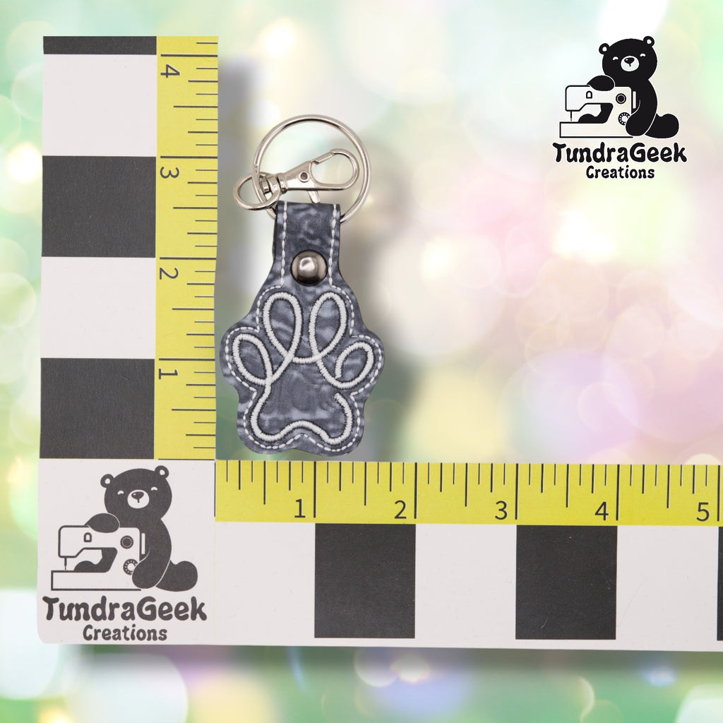 The Silver colorway paw print keychain shown against a black-and-white checkered background with yellow rulers for scale, measuring 1.5 inches wide by 3.5 inches tall including hardware. White embroidered paw print outline over marbled grey "Silver Fluidity" vinyl, matching grey tab, metal rivet closure, and silver split ring with swivel clip.