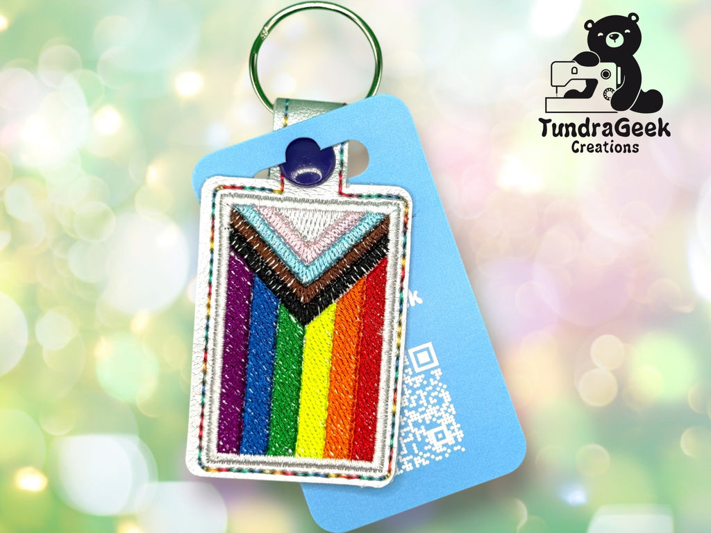 A keychain with a Progress Pride flag design is attached to a blue card displaying a QR code and the TundraGeek Creations logo, featuring a bear with a sewing machine.