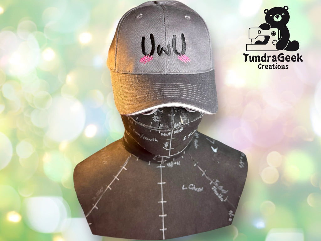 A gray baseball cap with UWU and pink blush marks is displayed on a black mannequin torso patterned with white markings. The TundraGeek Creations logo appears in the upper right corner.