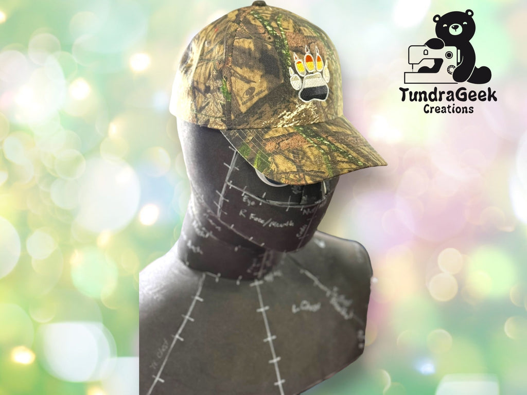 A camouflage baseball cap with a bear paw and rainbow claws sits on a mannequin bust. The TundraGeek Creations logo is in the upper right corner.