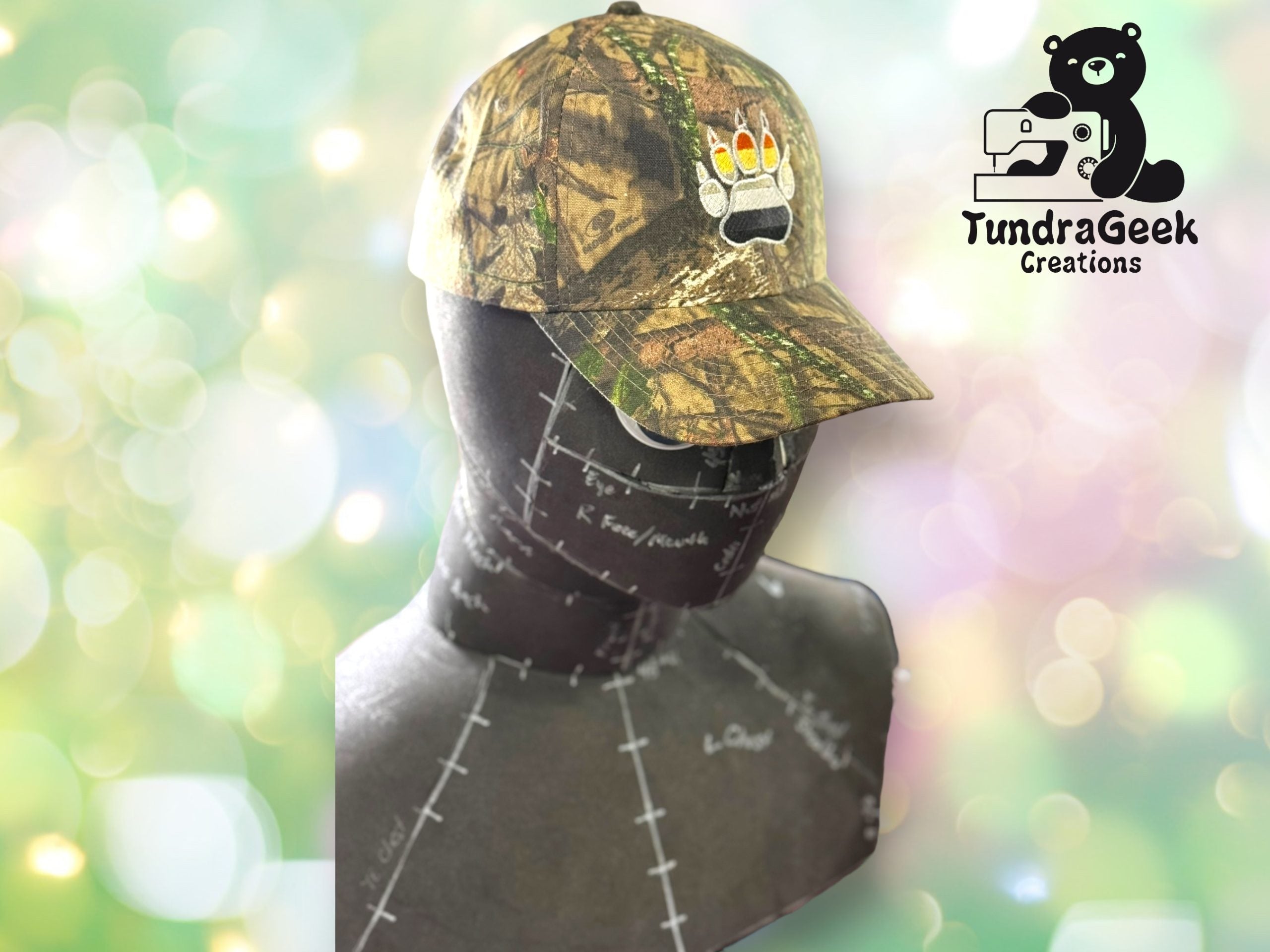 A camouflage baseball cap with a bear paw and rainbow claws sits on a mannequin bust. The TundraGeek Creations logo is in the upper right corner.